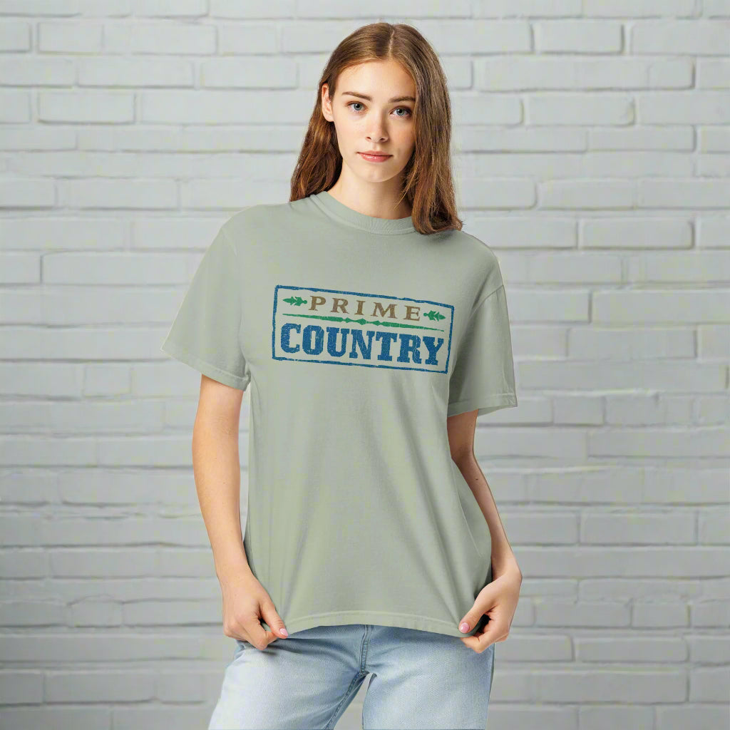A woman stands in front of a light gray brick wall, wearing a light sage green T-shirt with a large front print that reads 'PRIME COUNTRY' in rustic block letters. 'PRIME' appears in brown above the word 'COUNTRY' in faded blue, both enclosed in a distressed box with small green arrowhead graphics on either side of 'PRIME'.