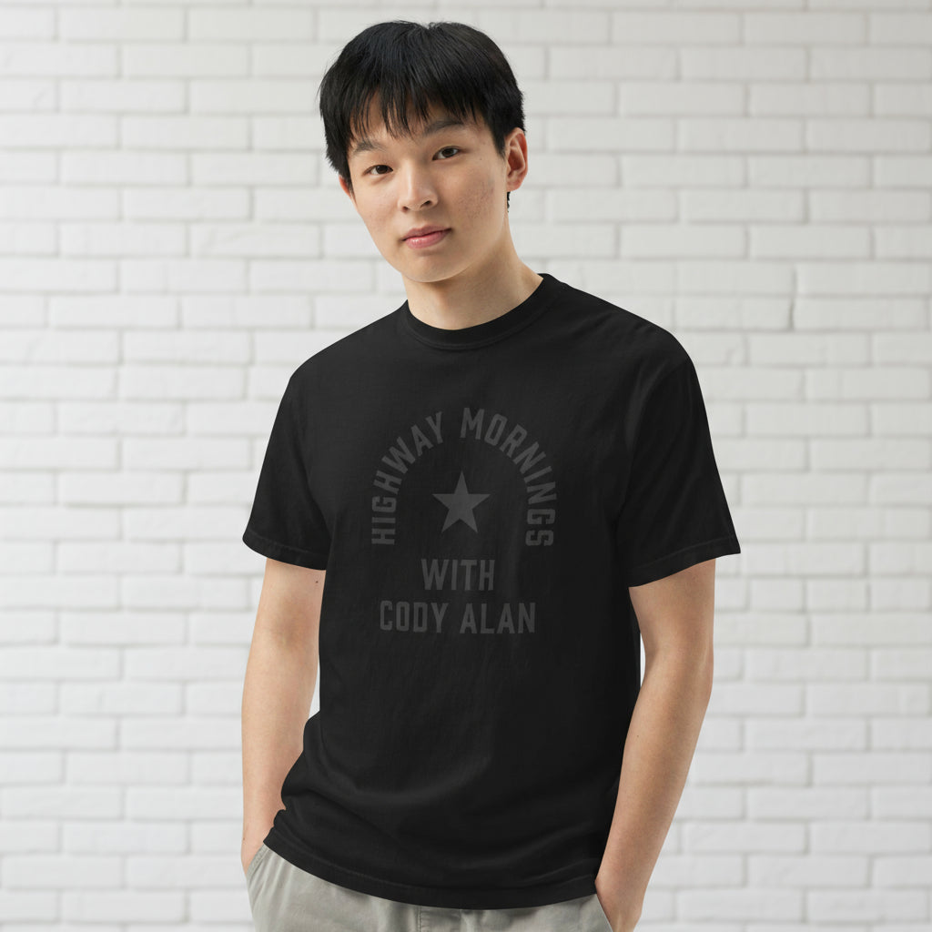 A man shown from the front wearing a black short-sleeve T-shirt with light gray pants, standing against a white brick background. The front of the shirt features a black-on-black design with a star in the center and the text 'HIGHWAY MORNINGS WITH CODY ALAN' printed in black above and below the star.
