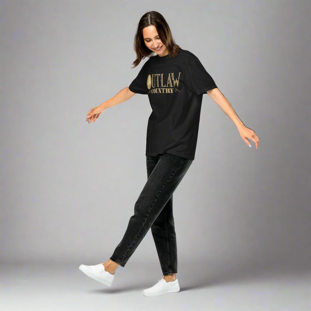Woman in motion wearing a black T-shirt with a large distressed gold oval emblem on the front featuring a silhouette of a cowboy on horseback, surrounded by vintage western-style text that reads 'OUTLAW COUNTRY'.