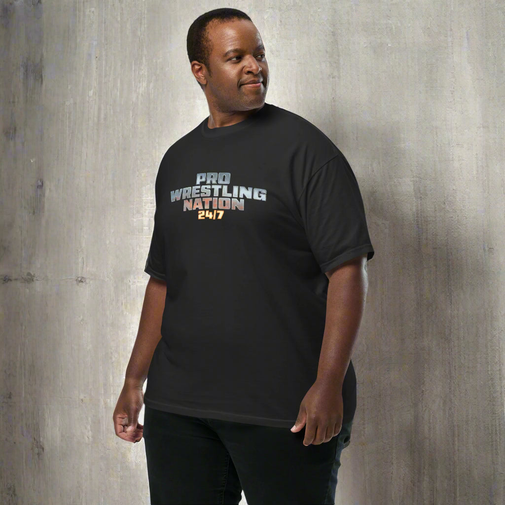 Side view of a man wearing a black 'Pro Wrestling Nation 24/7' graphic T-shirt, standing against a textured concrete wall.
