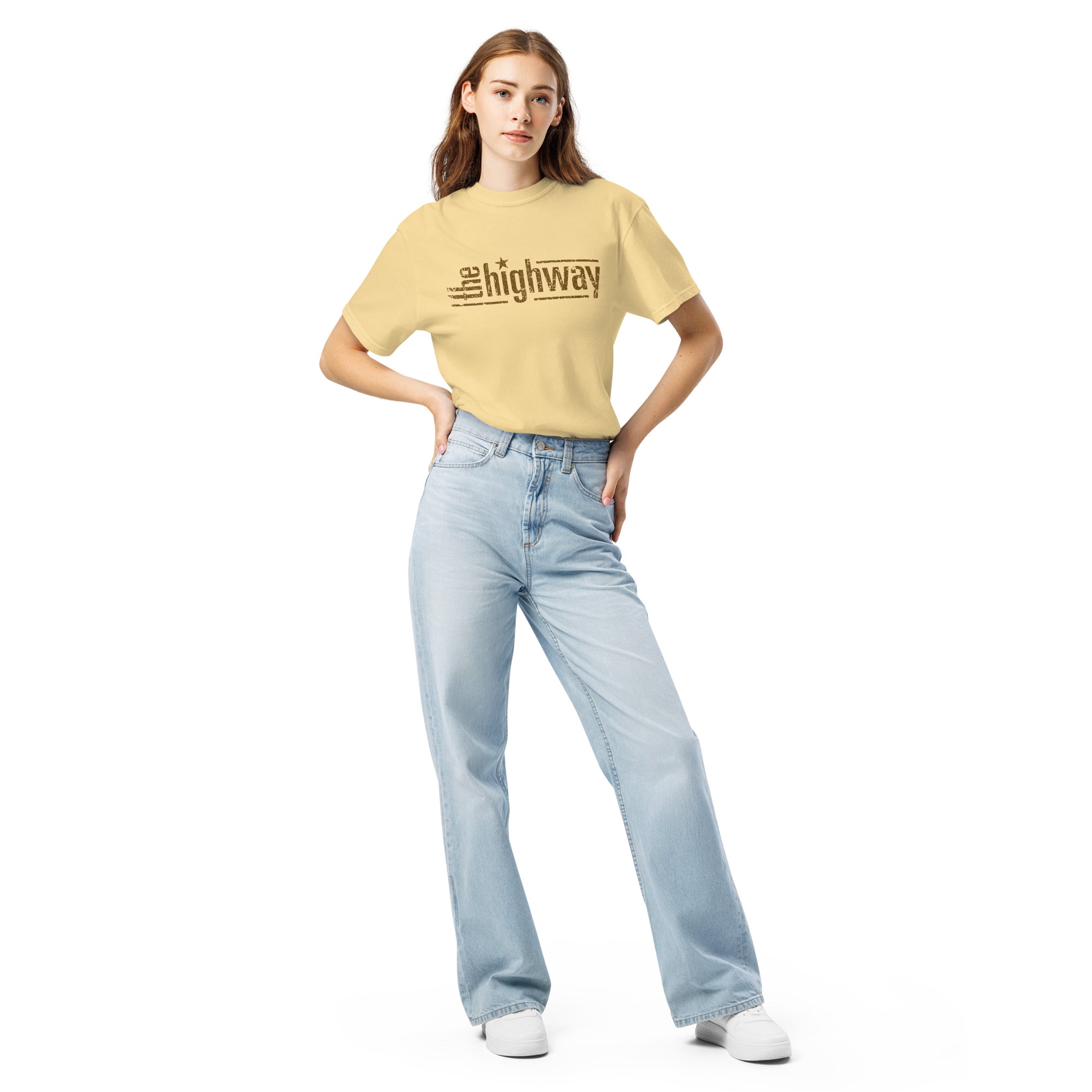 Woman standing in light-wash jeans and a pale yellow T-shirt with distressed brown text reading ‘the highway’, where the i in ‘highway’ is dotted with a small star.