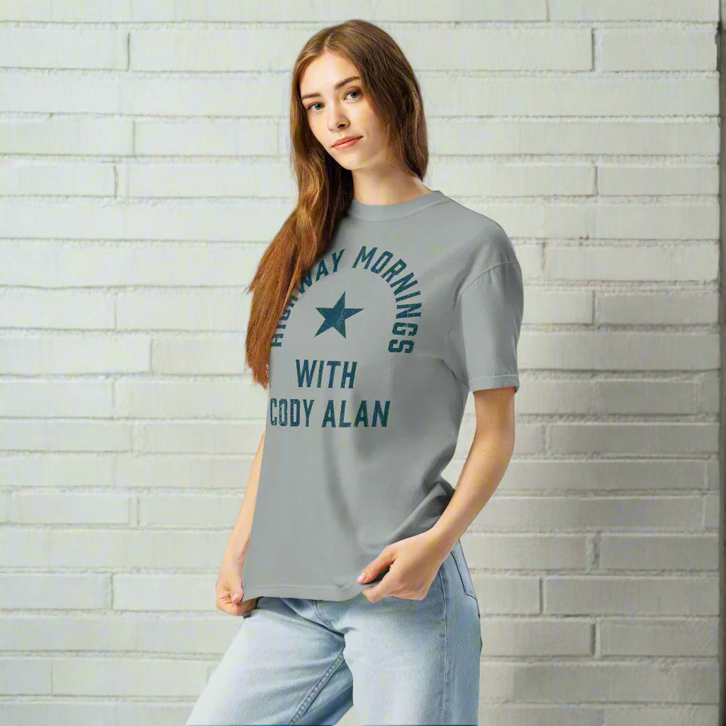 Woman standing against a white brick wall, wearing a gray short-sleeve t-shirt with distressed blue text 'HIGHWAY MORNINGS WITH CODY ALAN' arranged in an arc around a blue star on the chest.
