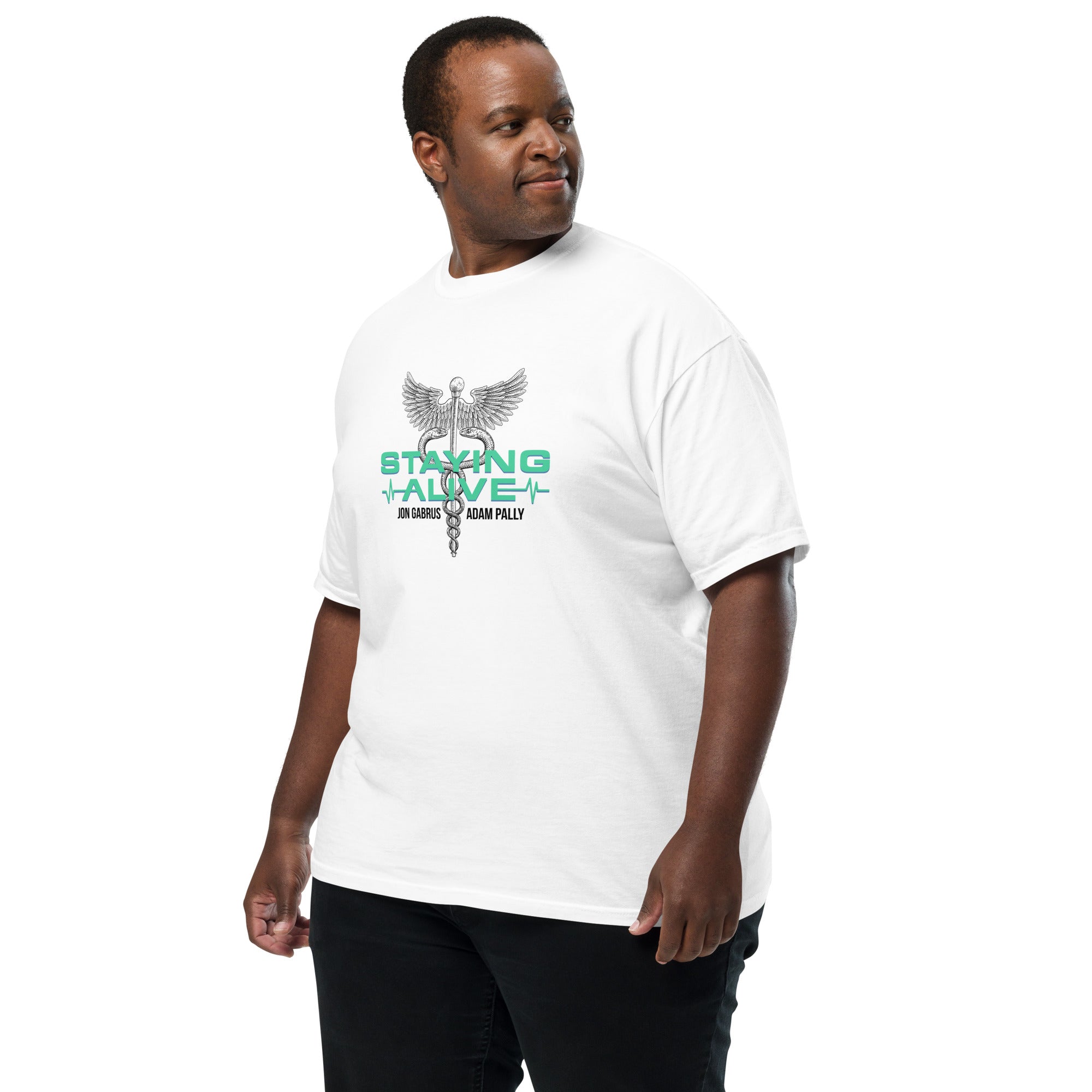 Man wearing a white graphic T-shirt featuring a caduceus (a winged staff with two snakes), mint green 'STAYING ALIVE' text, and black names 'Jon Gabrus' and 'Adam Pally' beneath an ECG line, paired with black pants.
