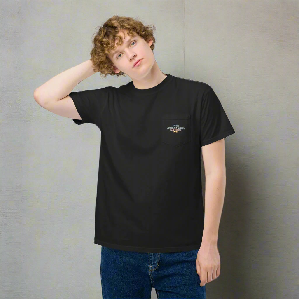 A young man wearing a black pocket t-shirt with the 'Pro Wrestling Nation 24/7' logo printed on the front pocket, standing in front of a white brick wall and resting one hand on the back of his head.
