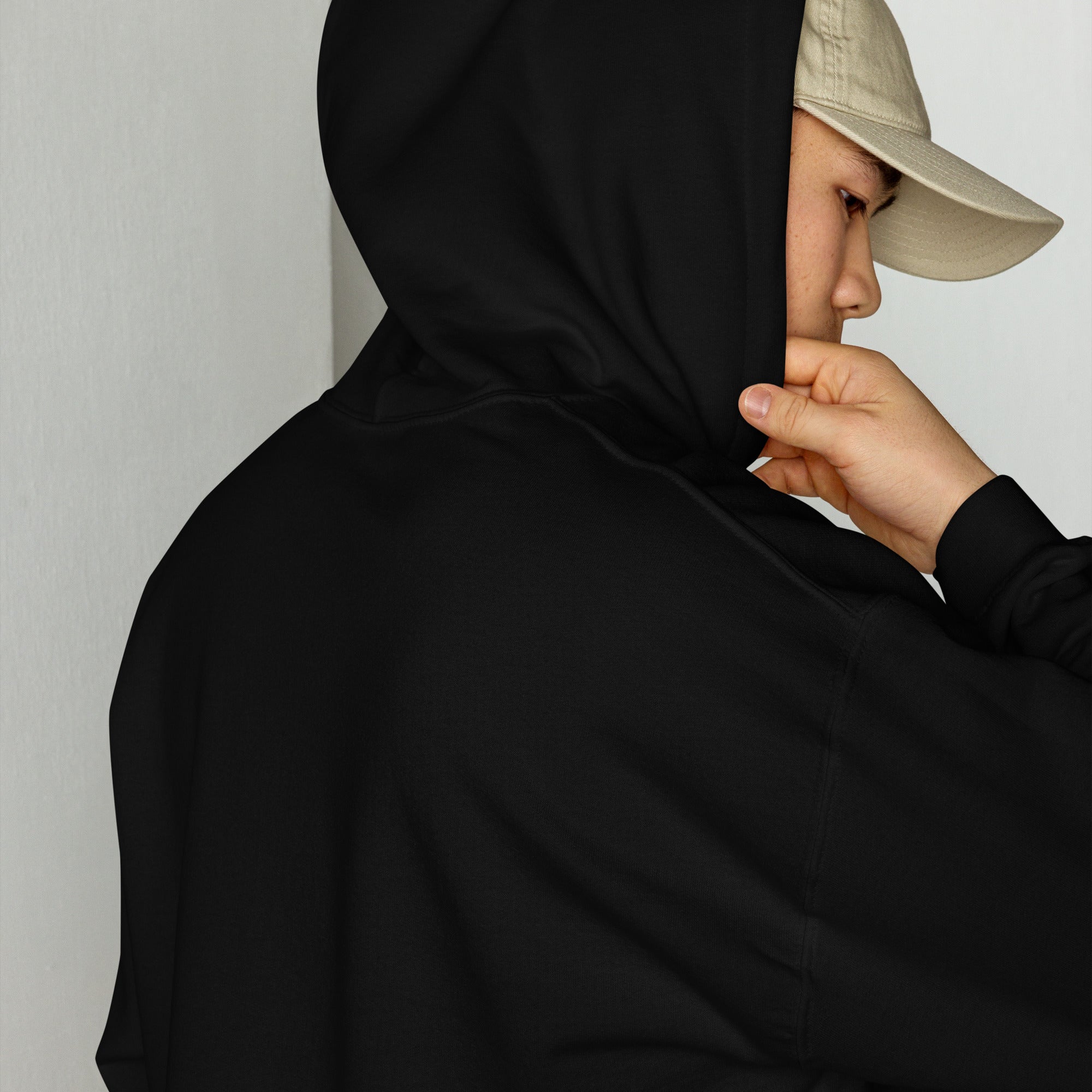 Close-up side profile of a person in a black Casuals with Katie Nolan hoodie, with the hood up and a beige cap worn underneath. No visible graphics from this angle.