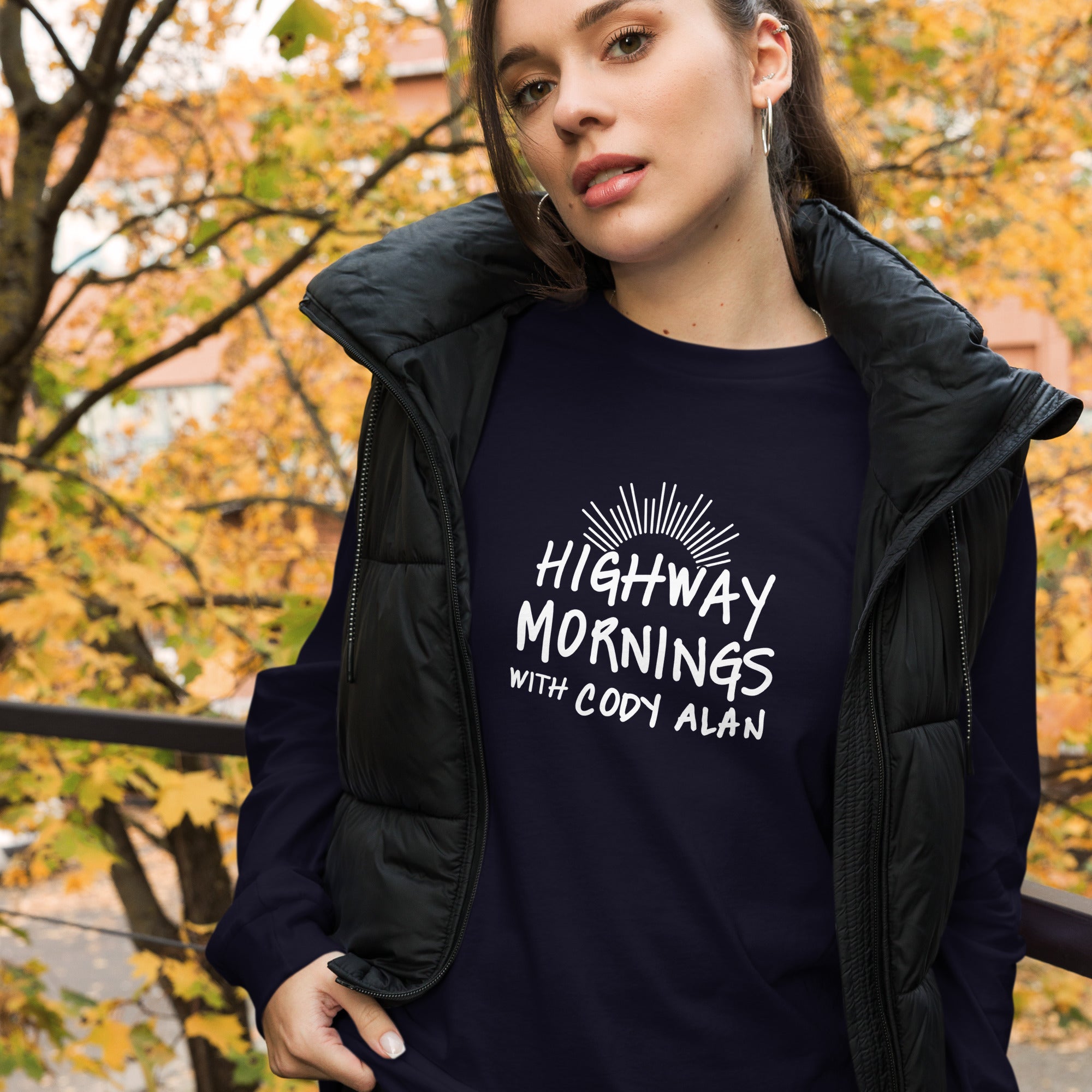 A woman standing outdoors during fall, wearing a navy long sleeve shirt with the phrase 'HIGHWAY MORNINGS WITH CODY ALAN' and a sunburst design in white. She’s layered with a black puffer vest.