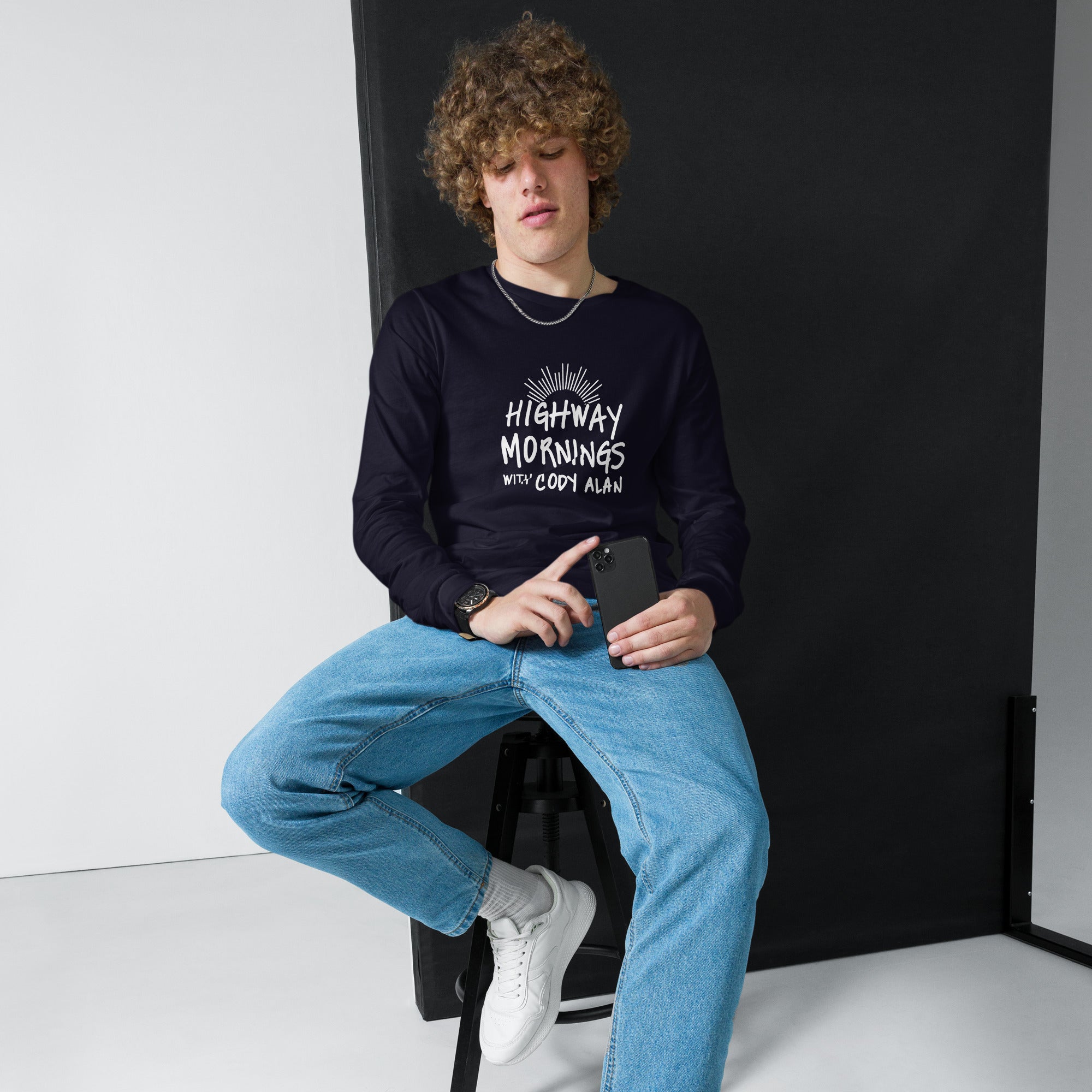 A young man sitting casually on a stool and holding a phone while wearing a navy long sleeve shirt with the 'HIGHWAY MORNINGS WITH CODY ALAN' graphic printed in white across the chest.