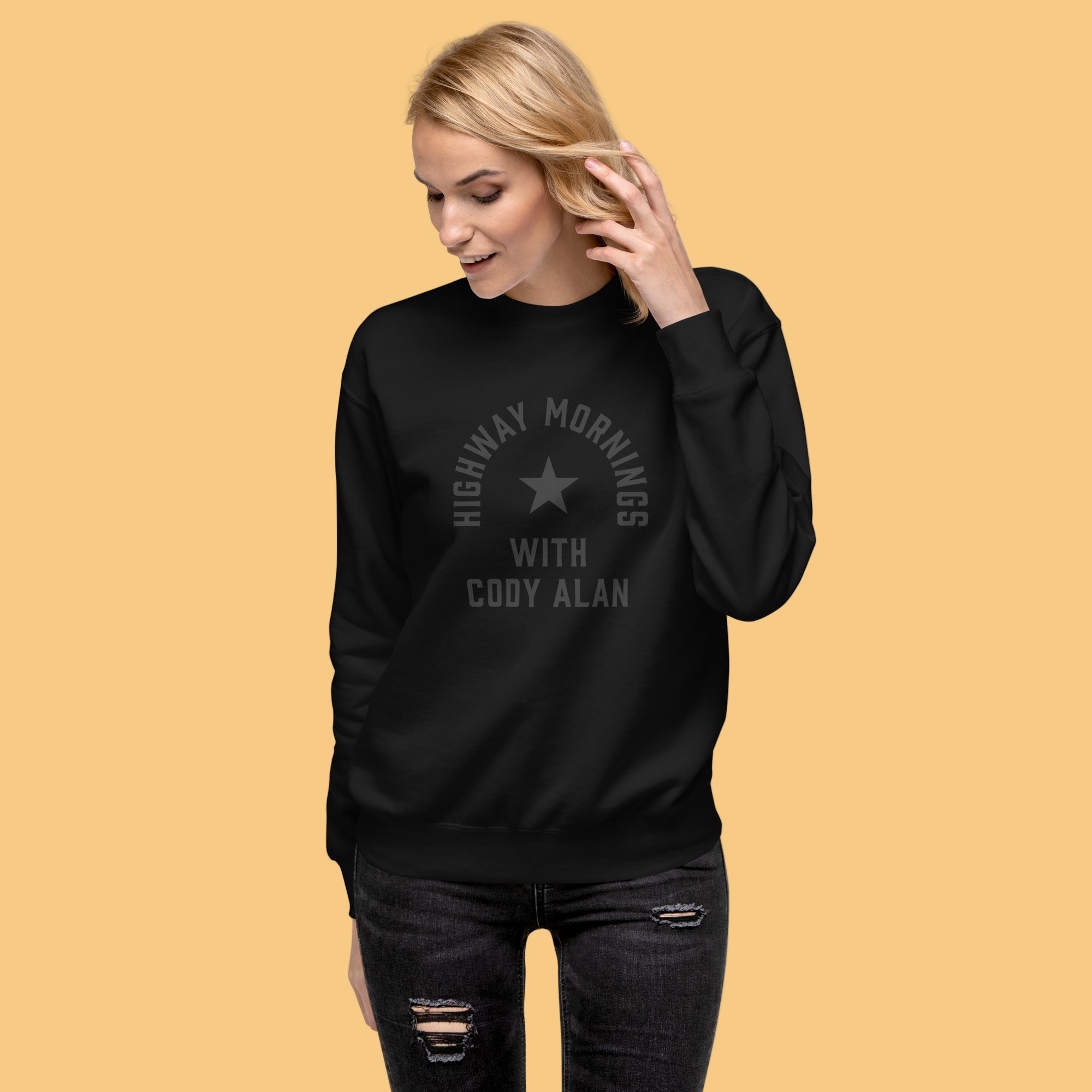 A woman shown from the front wearing a black crewneck sweatshirt with distressed black jeans. The sweatshirt design features a centered black star with the text 'HIGHWAY MORNINGS WITH CODY ALAN' printed in black above and below it in a tone-on-tone style.