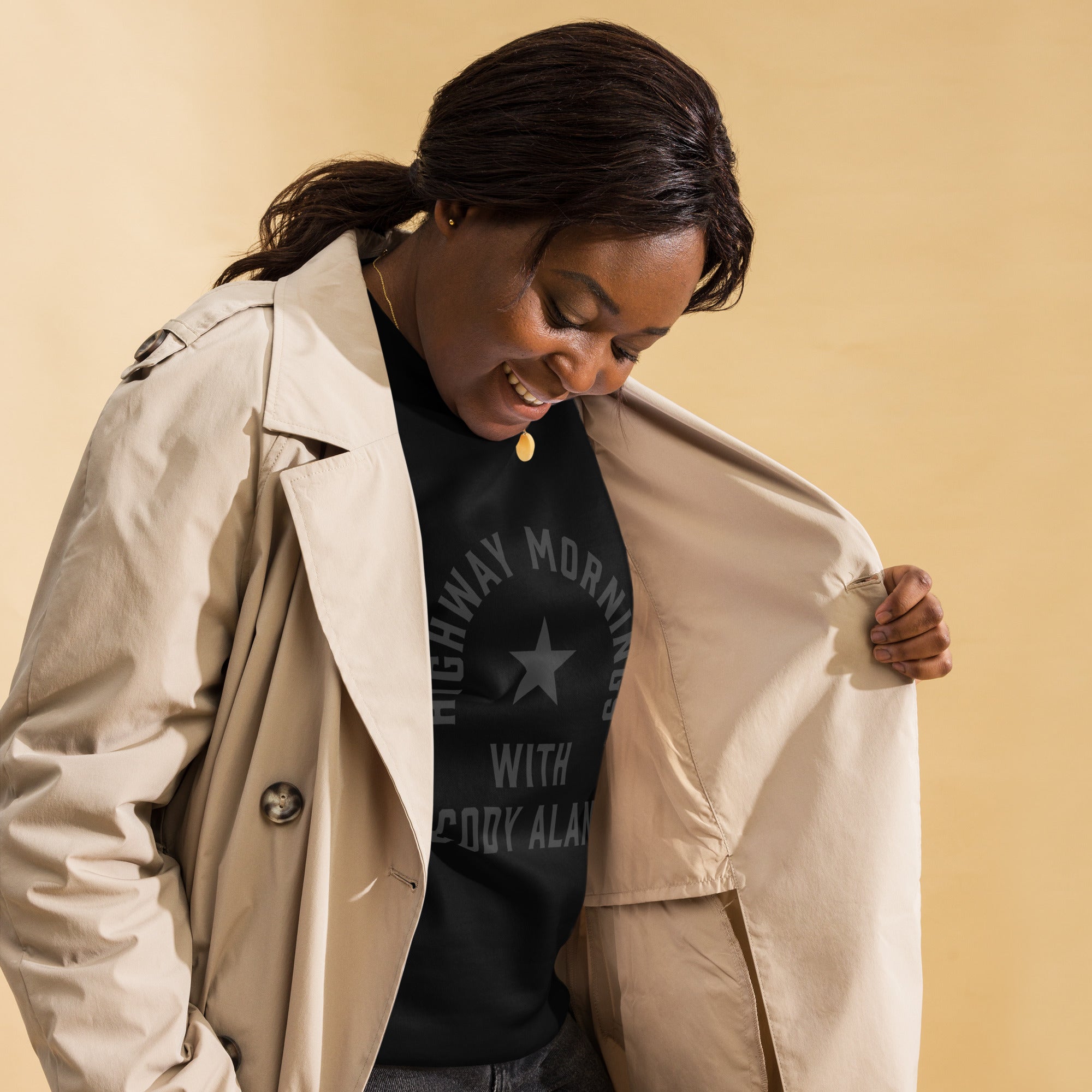 A woman shown in a right front view wearing a black crewneck sweatshirt under a light beige trench coat. She is smiling and opening her coat to reveal the sweatshirt’s front design, which features a star and the text 'HIGHWAY MORNINGS WITH CODY ALAN' printed in black for a subtle tone-on-tone look.