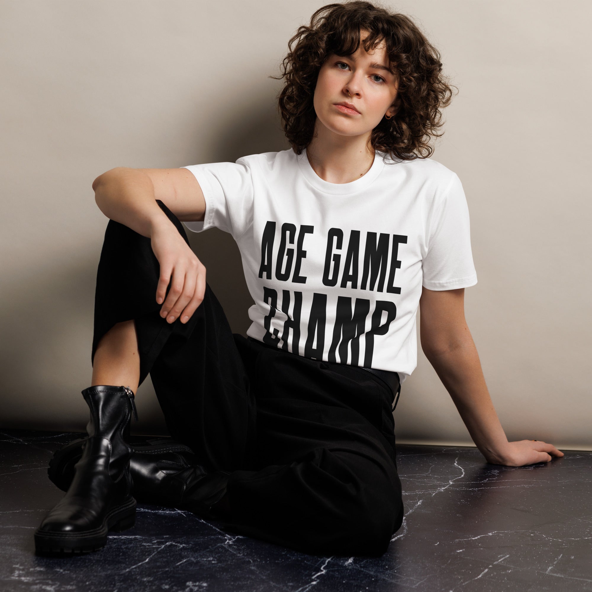 Woman sitting on the floor wearing a white T-shirt with bold black text 'AGE GAME CHAMP', paired with black pants and black boots.