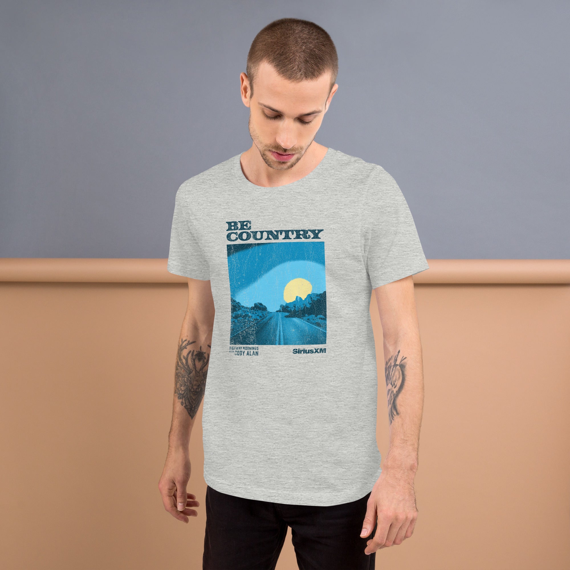 Man standing in a gray t-shirt printed with a cracked blue and yellow graphic of a desert road at sunrise, with 'BE COUNTRY' across the top and 'HIGHWAY MORNINGS WITH CODY ALAN' and 'SiriusXM' printed below.