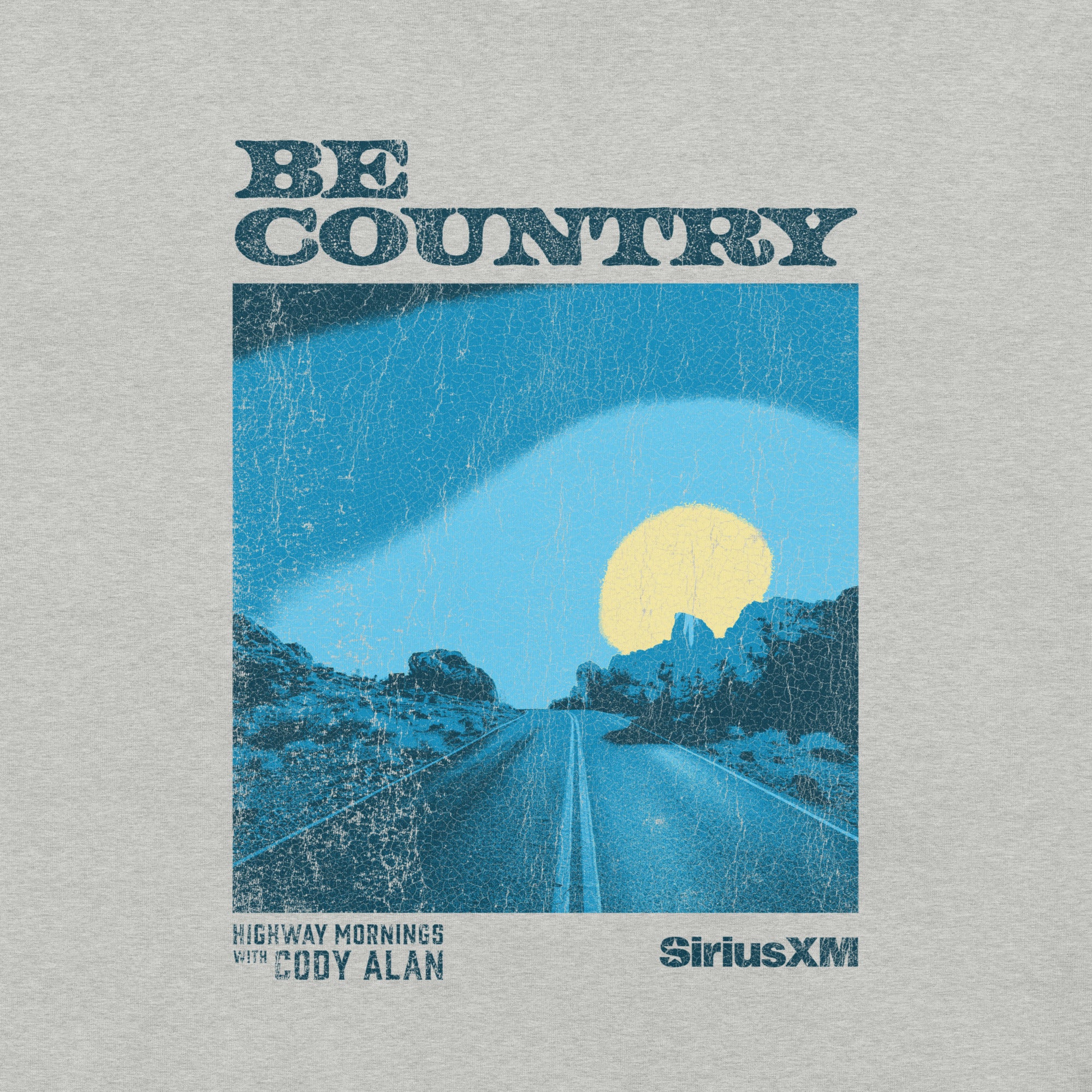 Close-up of a textured print showing a two-lane road through rocky terrain under a blue sky with a yellow sun. Text includes 'BE COUNTRY', 'HIGHWAY MORNINGS WITH CODY ALAN', and 'SiriusXM'.