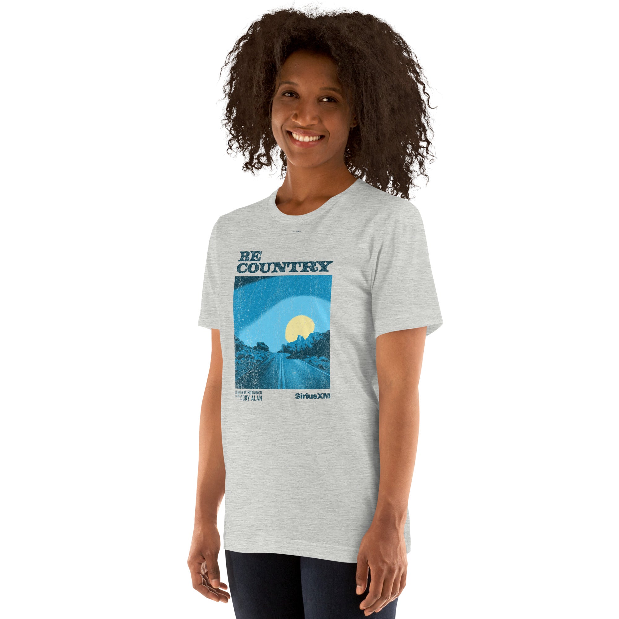 Woman smiling and wearing a heather gray t-shirt printed with a cracked blue and yellow graphic of a desert sunrise and the words 'BE COUNTRY', 'HIGHWAY MORNINGS WITH CODY ALAN', and 'SiriusXM'.