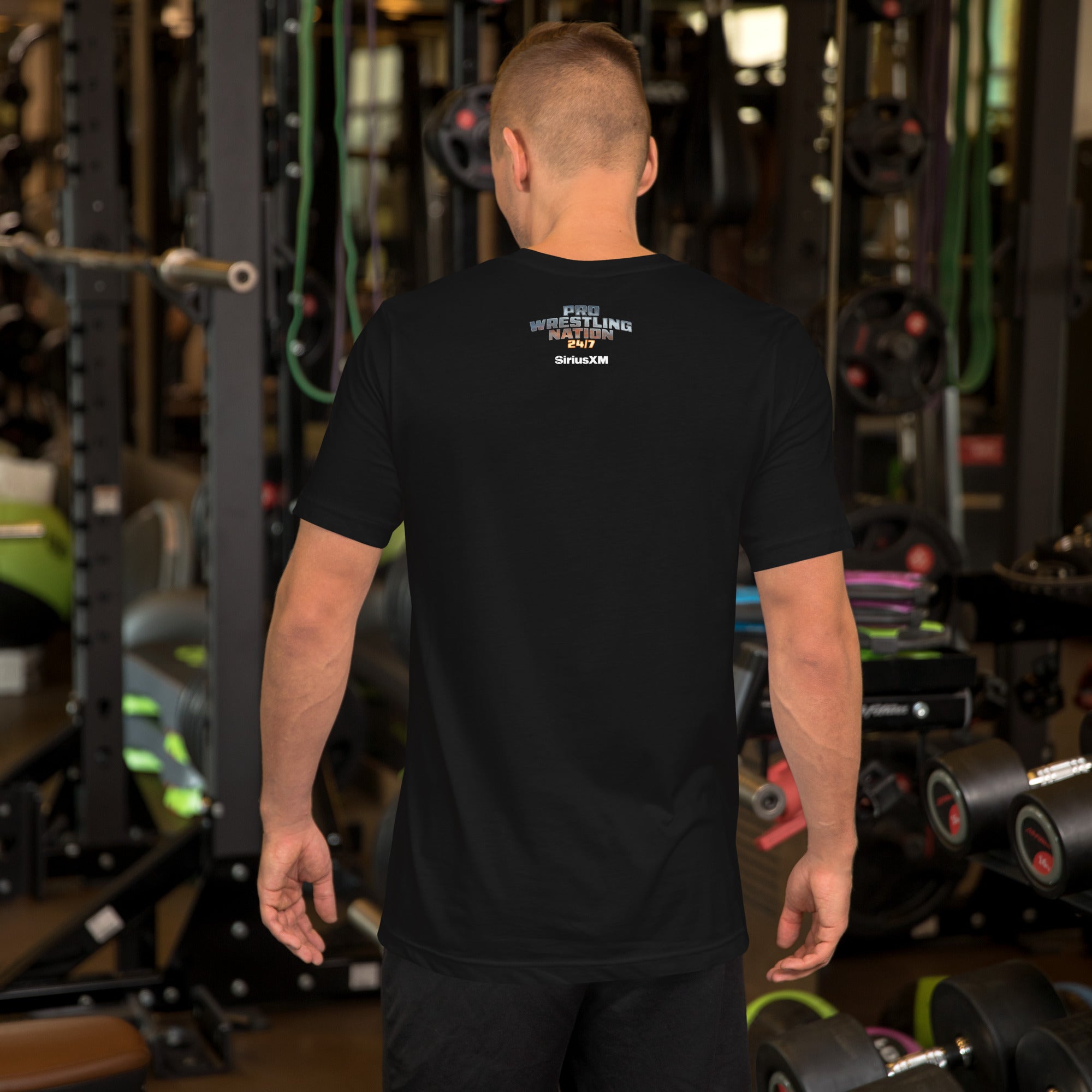 A man in a gym facing away from the camera wearing a black T-shirt with a small back print that reads 'PRO WRESTLING NATION 24/7' and 'SiriusXM' in metallic and white text.