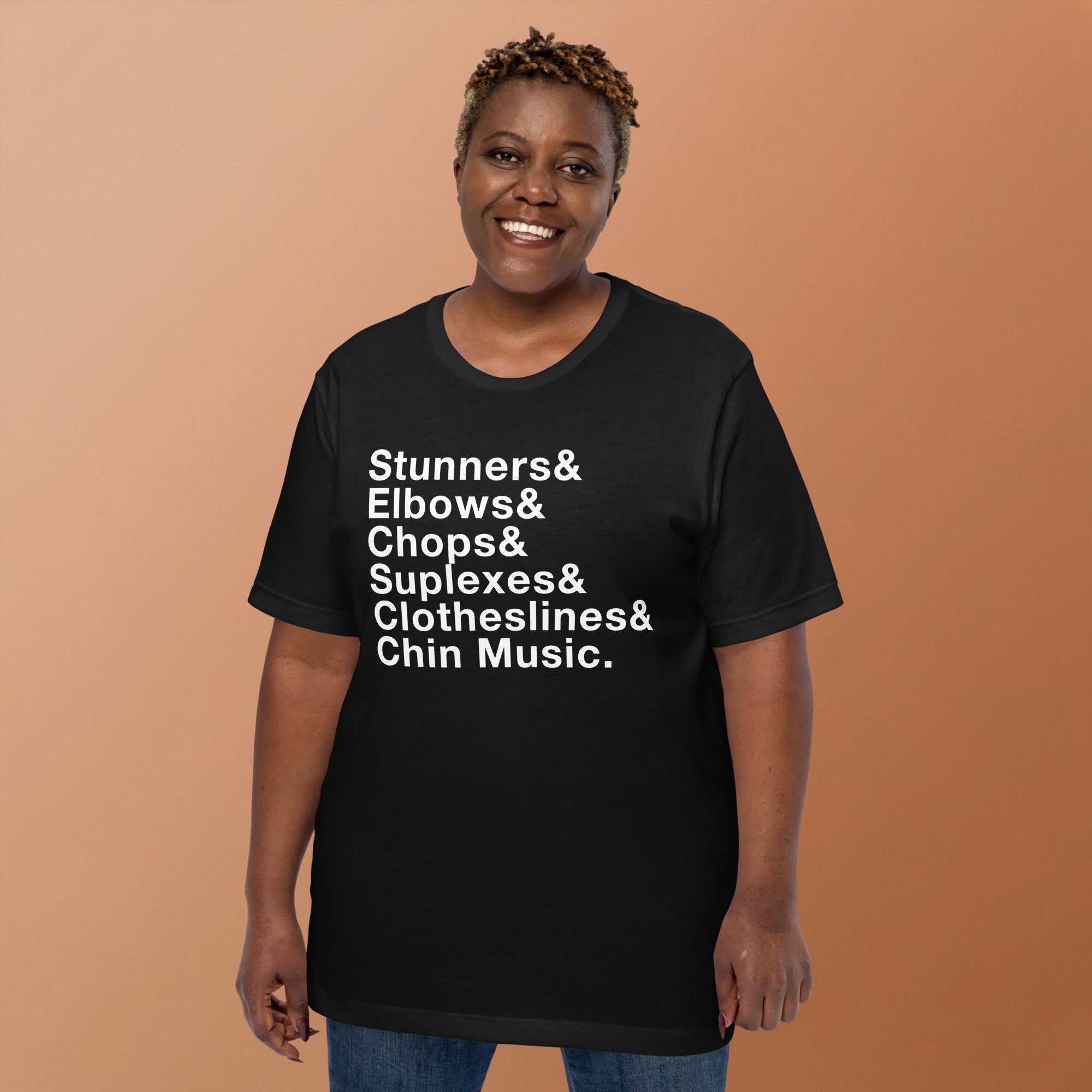 A smiling woman standing against a warm beige backdrop wearing a black T-shirt with bold white stacked text that reads: 'Stunners& Elbows& Chops& Suplexes& Clotheslines& Chin Music.'