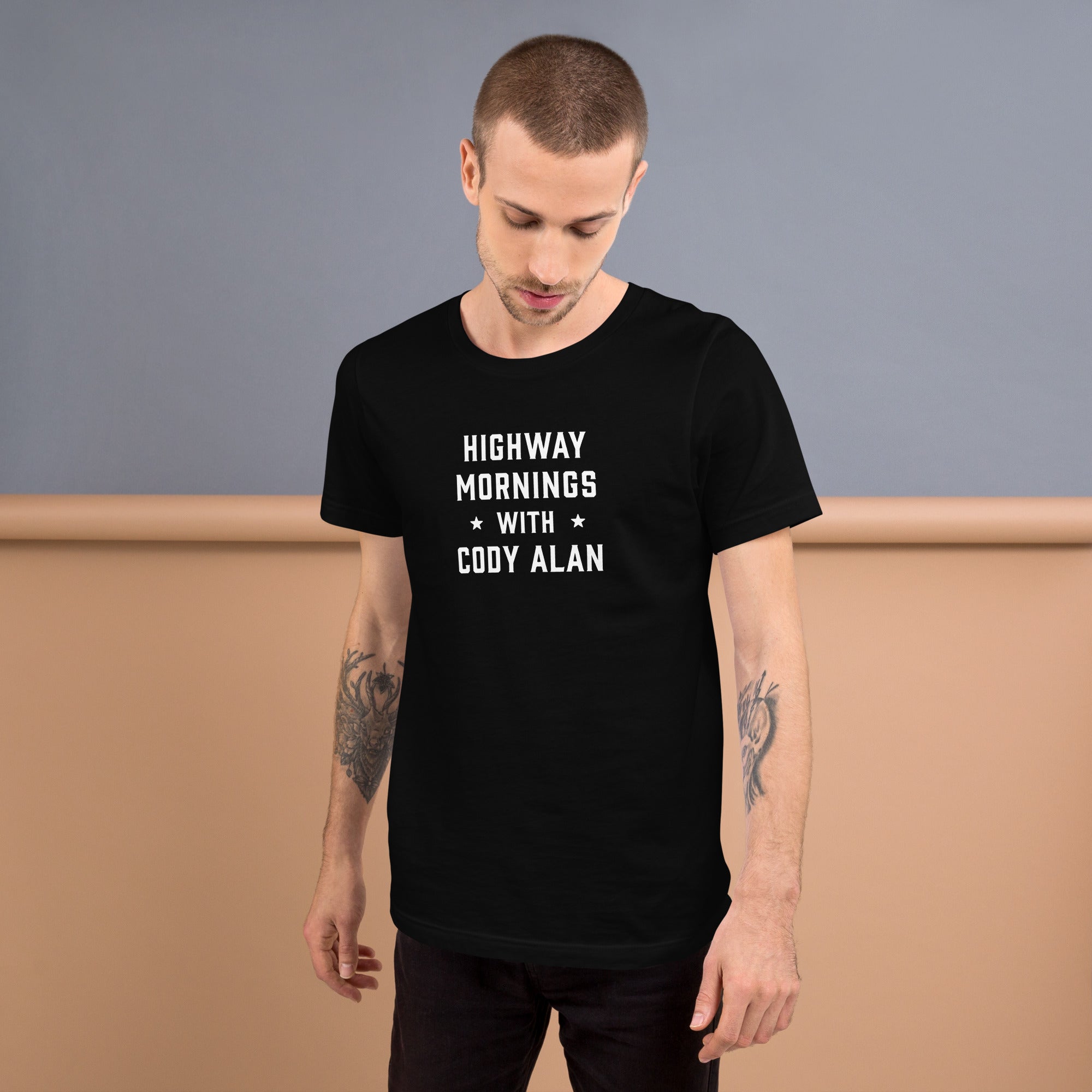 A man looking down while wearing a black t-shirt with the phrase 'HIGHWAY MORNINGS WITH CODY ALAN' printed in white block letters, styled with black jeans and tattoos visible on both arms.