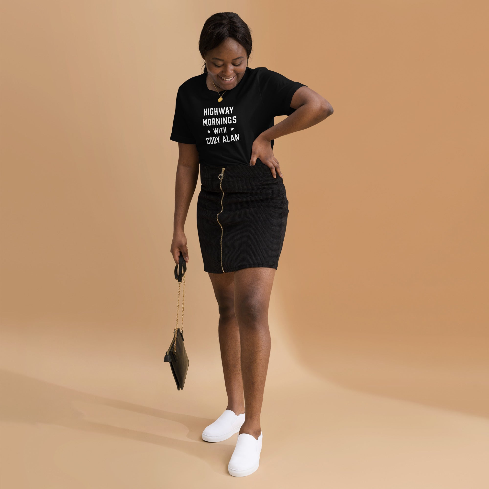 A smiling woman wearing a black t-shirt with white 'HIGHWAY MORNINGS WITH CODY ALAN' text, paired with a black zip-up skirt and white slip-on shoes.