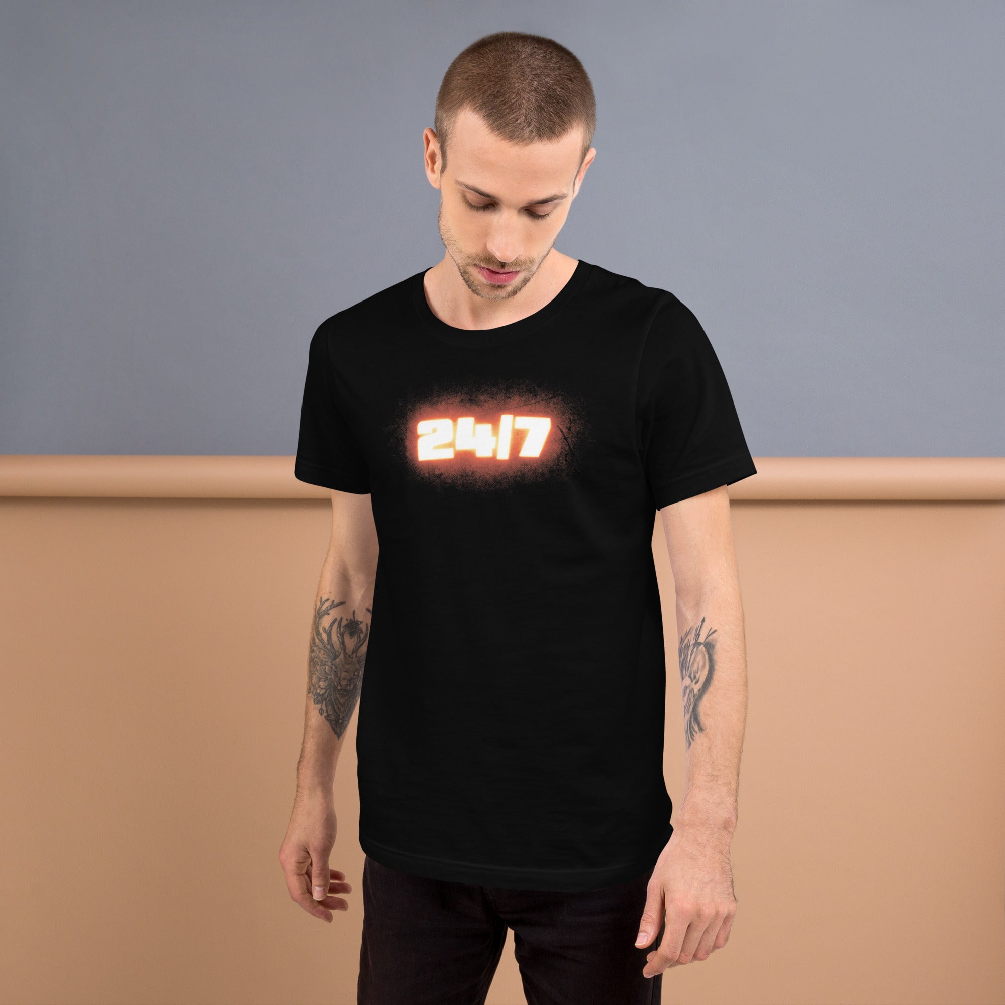 Man standing indoors wearing a black t-shirt featuring the glowing orange ‘24/7’ graphic centered on the chest, looking down with hands at his sides.