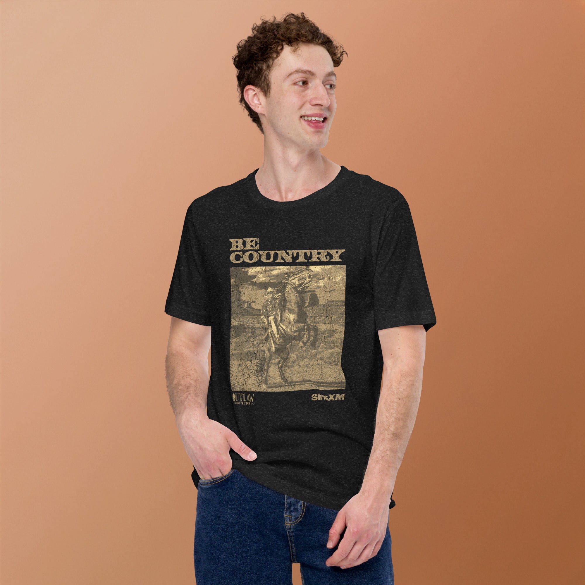 Front view of a man wearing a black heather t-shirt with a sepia-toned graphic that says 'BE COUNTRY' above a cowboy riding a horse. Below the image, it reads 'OUTLAW COUNTRY' and 'SiriusXM'.