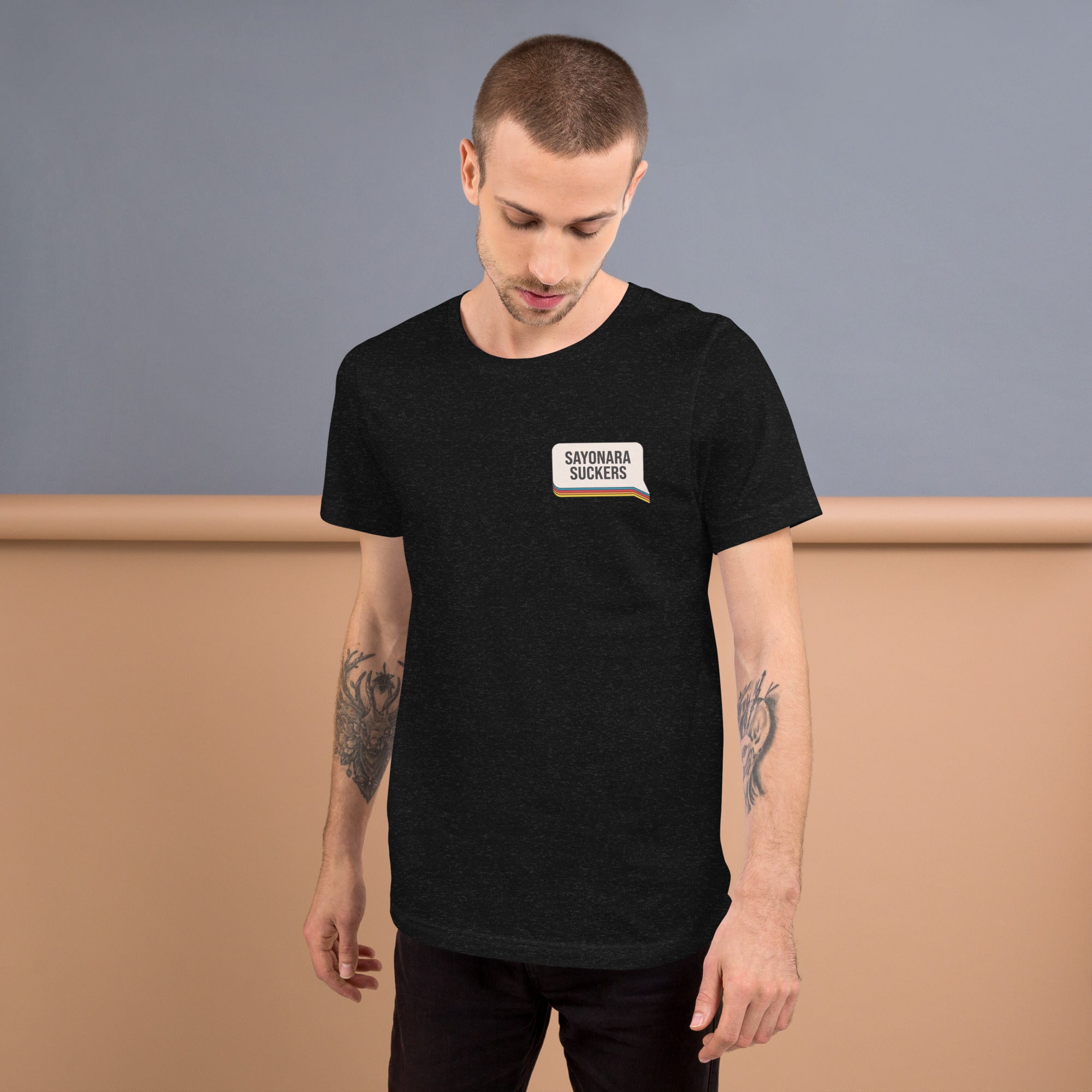 A man with arm tattoos wearing a black heather T-shirt featuring a chest graphic that reads 'SAYONARA SUCKERS' with a small rainbow stripe underneath.