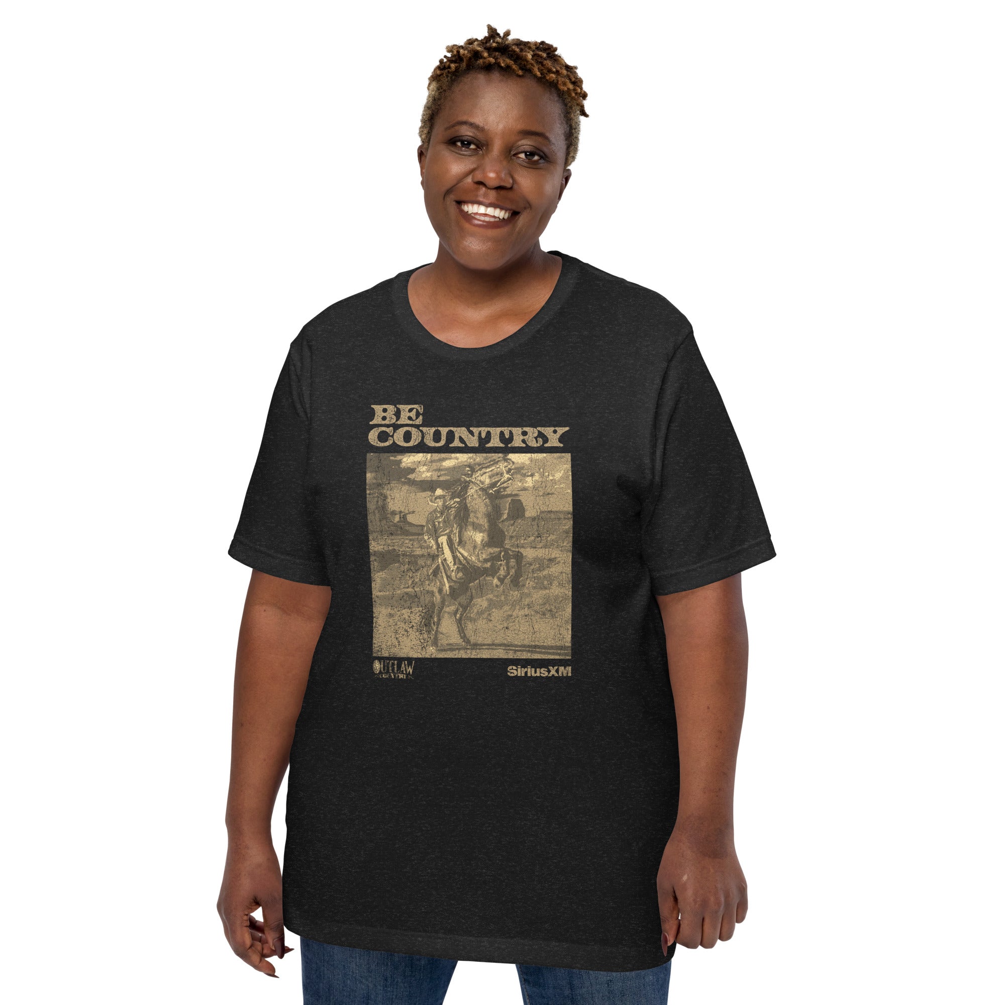 A woman wears a black heather t-shirt with a tan graphic of a cowboy on a rearing horse. The shirt reads 'BE COUNTRY' across the top, with 'OUTLAW COUNTRY' and 'SiriusXM' logos below the image.