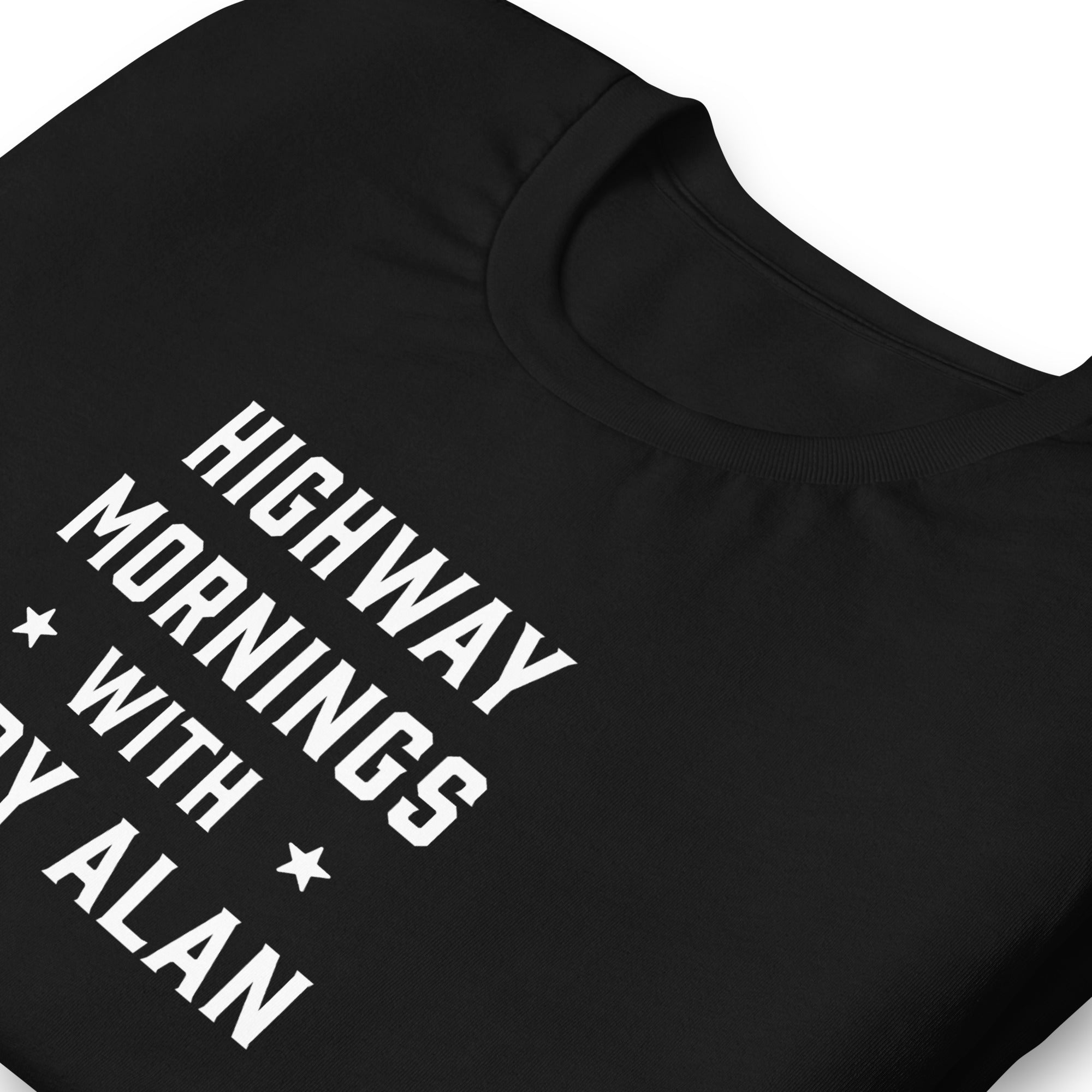A close-up of a folded black t-shirt showing the white printed text 'HIGHWAY MORNINGS WITH CODY ALAN' in bold block letters, with a star on each side of 'WITH'.