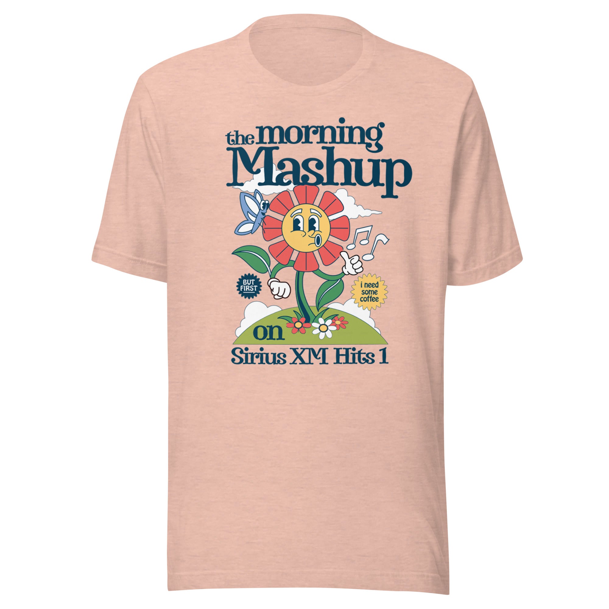 Peach-colored unisex T-shirt featuring a cartoon flower character with text reading 'the morning Mashup on Sirius XM Hits 1.' The flower holds a sign that says 'i need some coffee,' with musical notes, clouds, a butterfly, and a hill in the background.