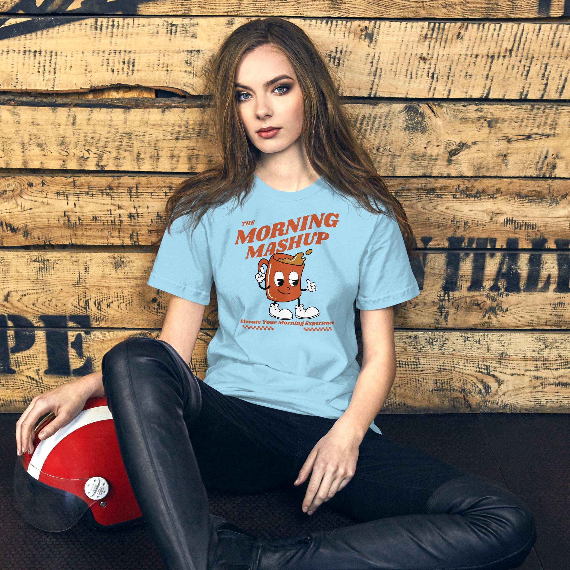 Woman with long brown hair sitting against a rustic wood wall, wearing a light blue T-shirt featuring a cartoon coffee mug with eyes, sneakers, and arms pointing outward. The mug has coffee splashing out of it, with the words 'THE MORNING MASHUP' above and 'Elevate Your Morning Experience' below in bold orange text and checkerboard accents.