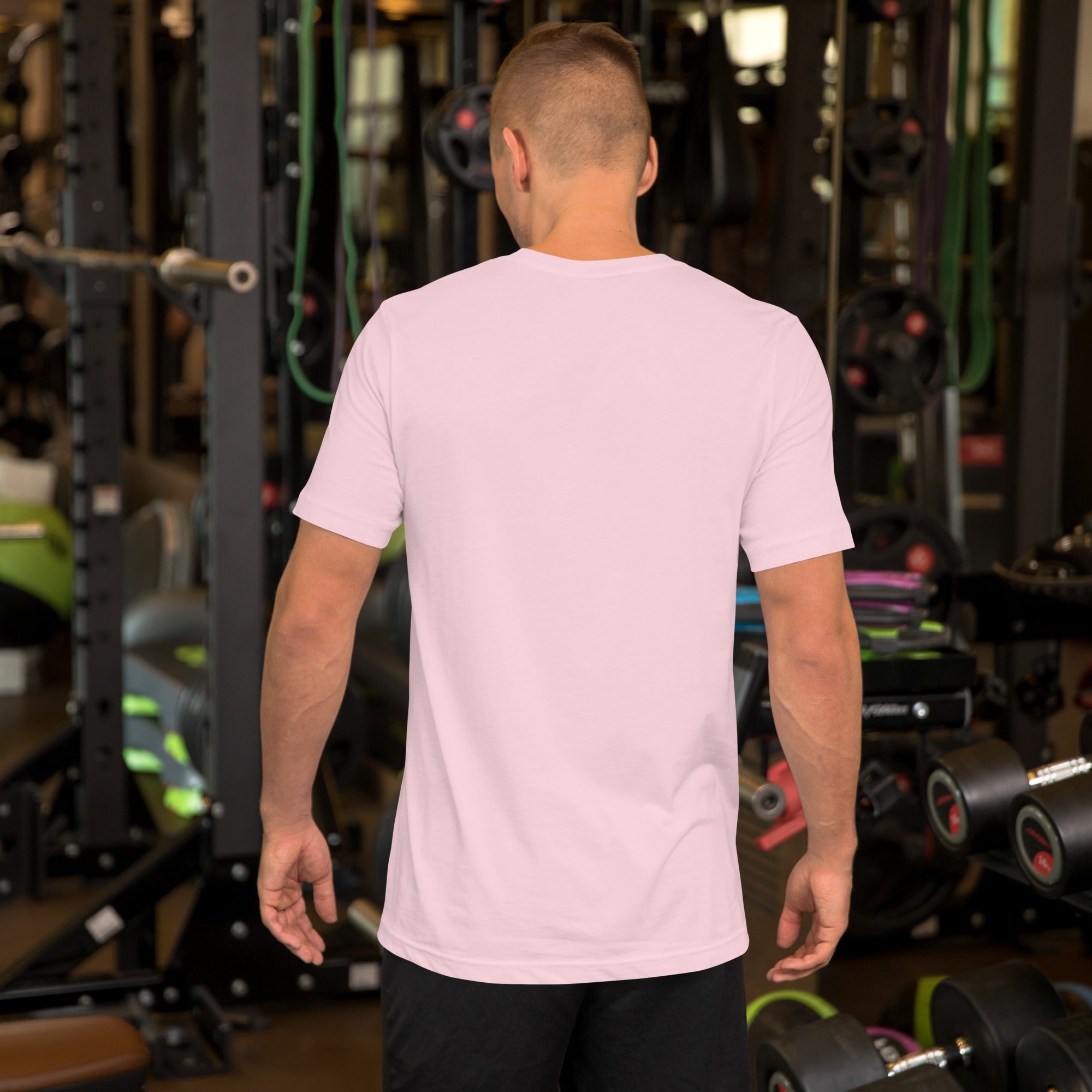 A man standing in a gym wearing a light pink short-sleeve t-shirt, shown from the back, surrounded by fitness equipment and weights.