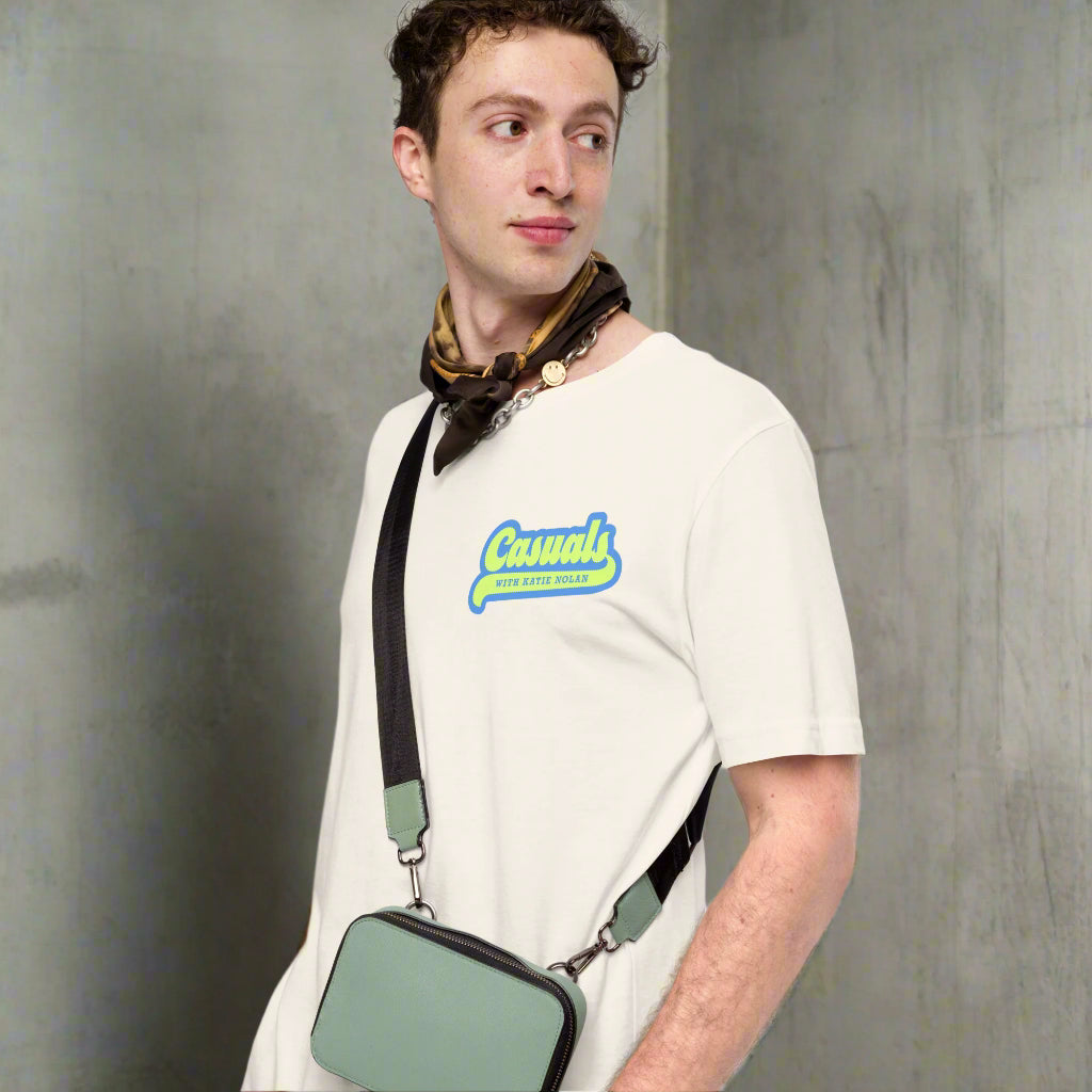 A man wearing a vintage white T-shirt styled with a brown patterned scarf and a green crossbody bag; the shirt displays a graphic on the left chest with the word 'Casuals' in bold blue font and 'WITH KATIE NOLAN' in yellow underneath.