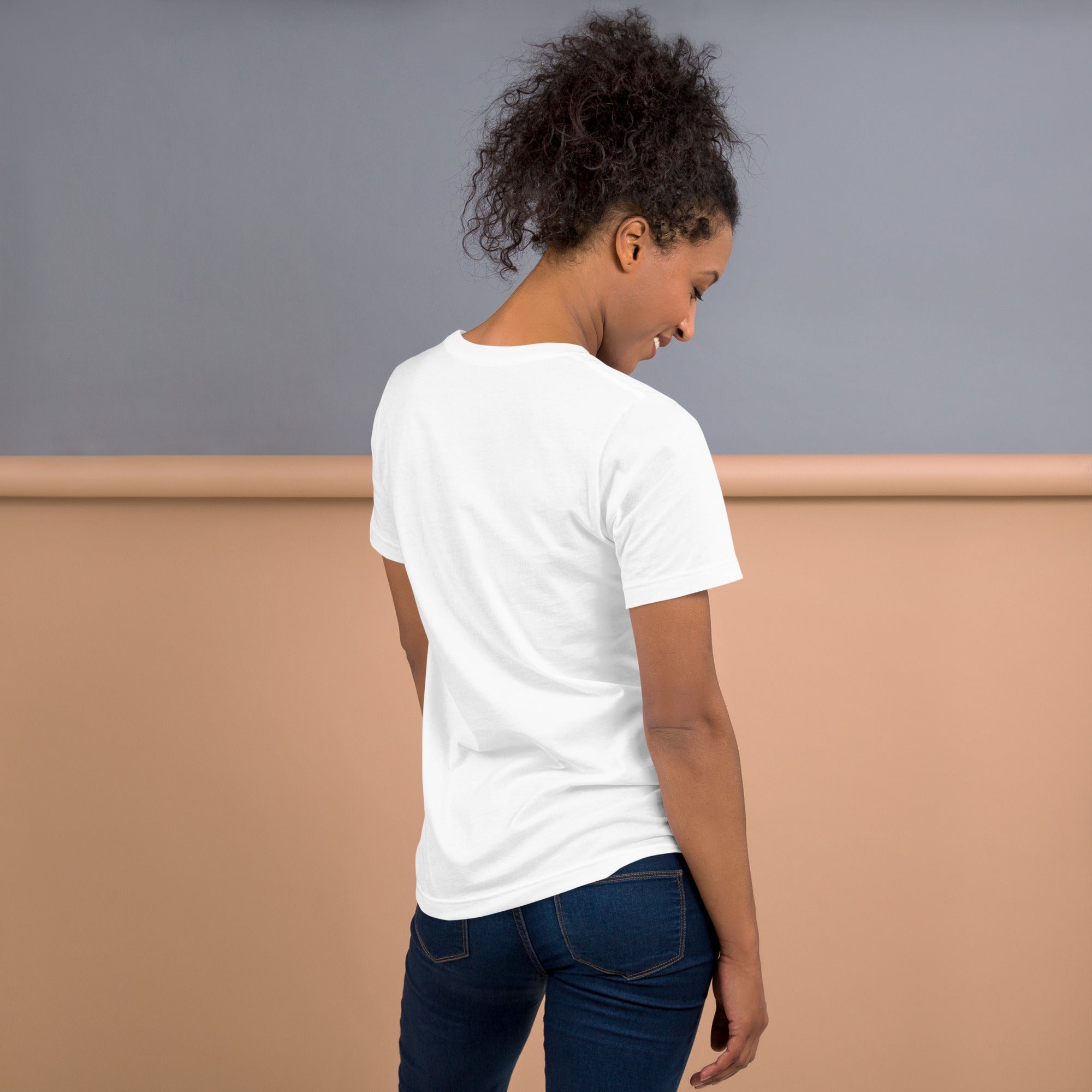 Woman wearing a plain white unisex T-shirt viewed from the back, standing with head turned slightly and arms behind her back.
