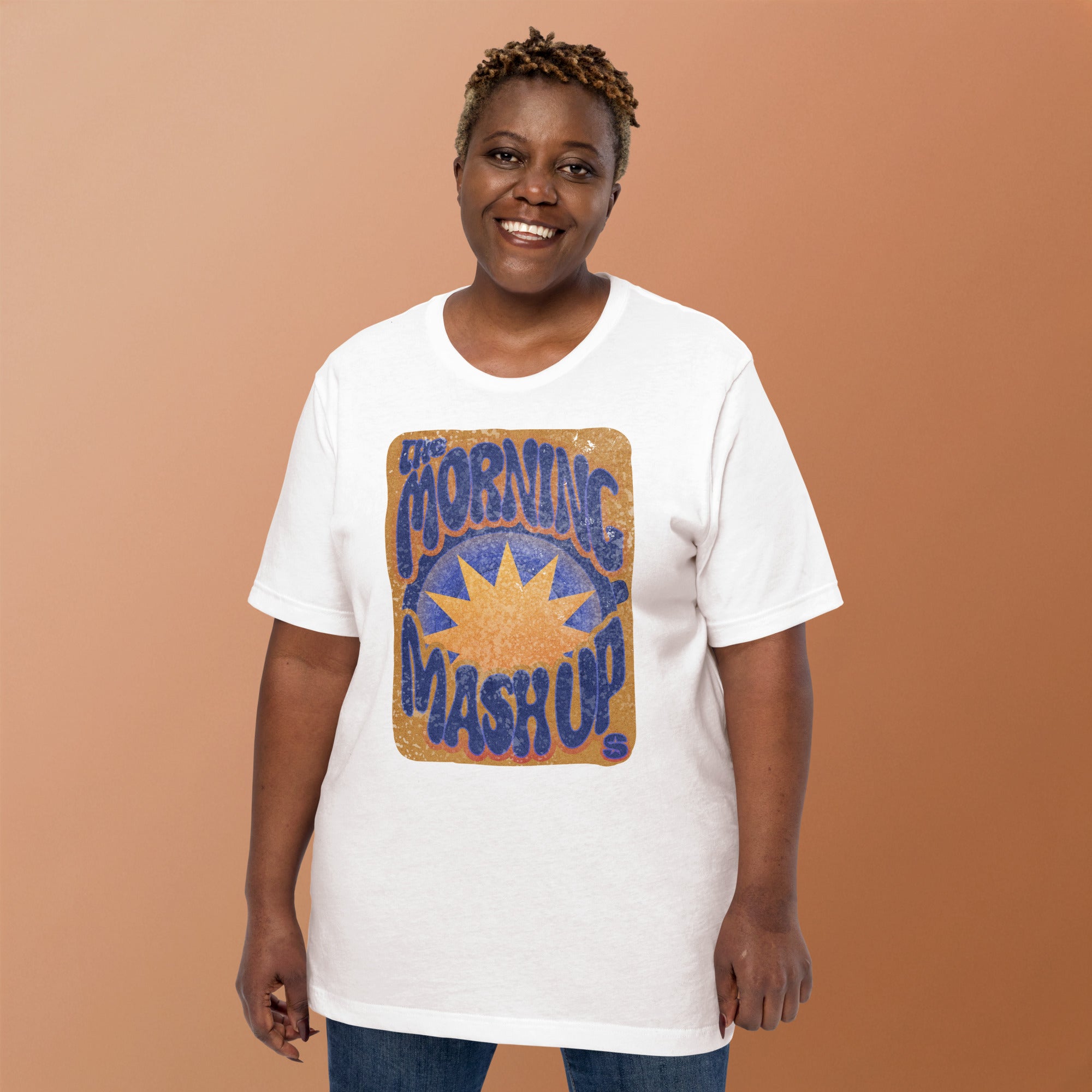 A woman smiling while wearing a white short-sleeve T-shirt with a large distressed retro graphic on the front. The design features the words 'The Morning Mash Up' in wavy blue letters surrounding an orange sunburst on a brown background. The SiriusXM 'S-Star' logo appears in the bottom right corner of the graphic.