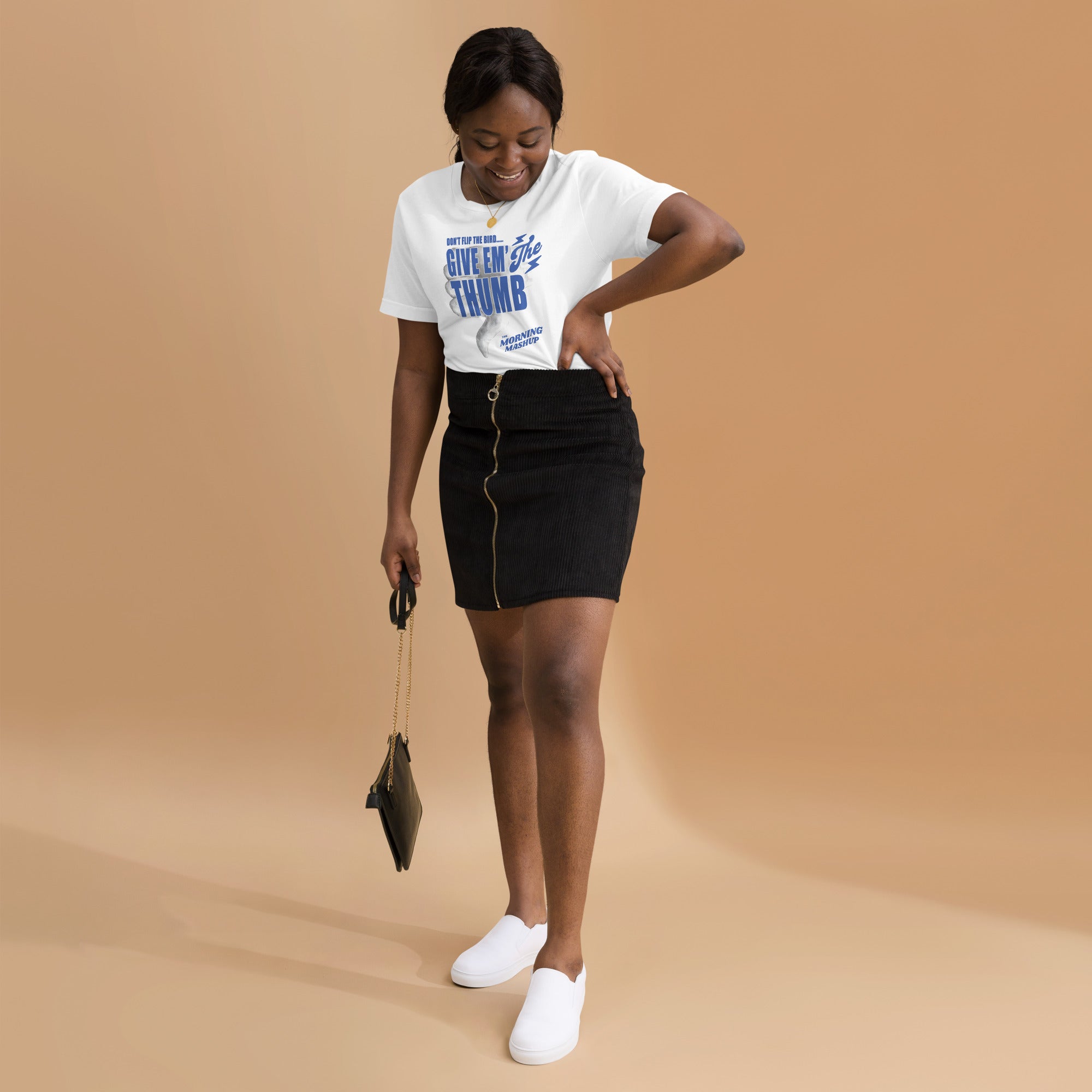 Woman wearing a white T-shirt with the 'GIVE EM’ THE THUMB' design and a black zippered skirt, smiling and looking down with one hand on her hip.