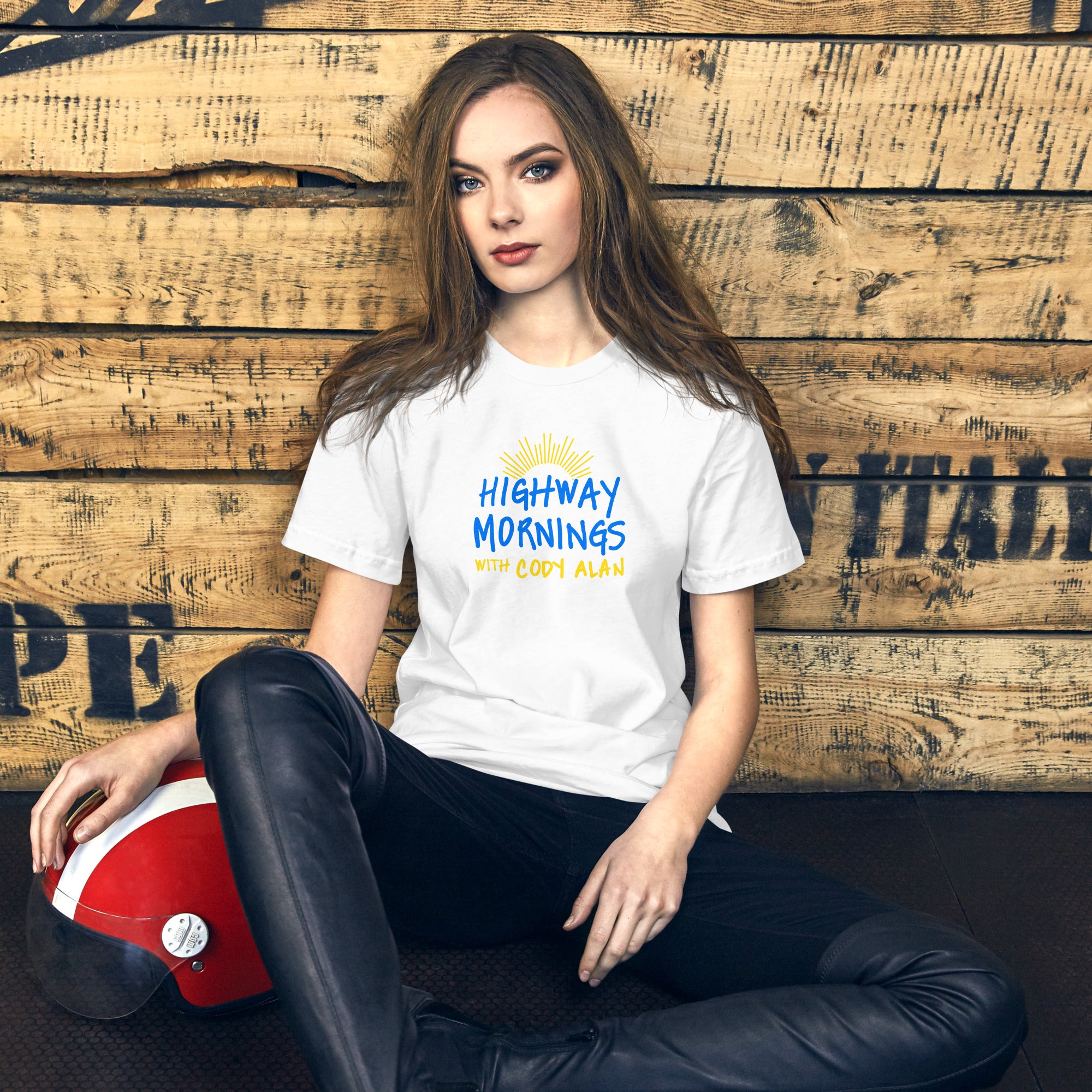 A woman seated in front of a rustic wood wall, wearing a white t-shirt with the phrase 'HIGHWAY MORNINGS WITH CODY ALAN' printed in blue and yellow beneath a sunburst graphic. A red and white helmet rests beside her.