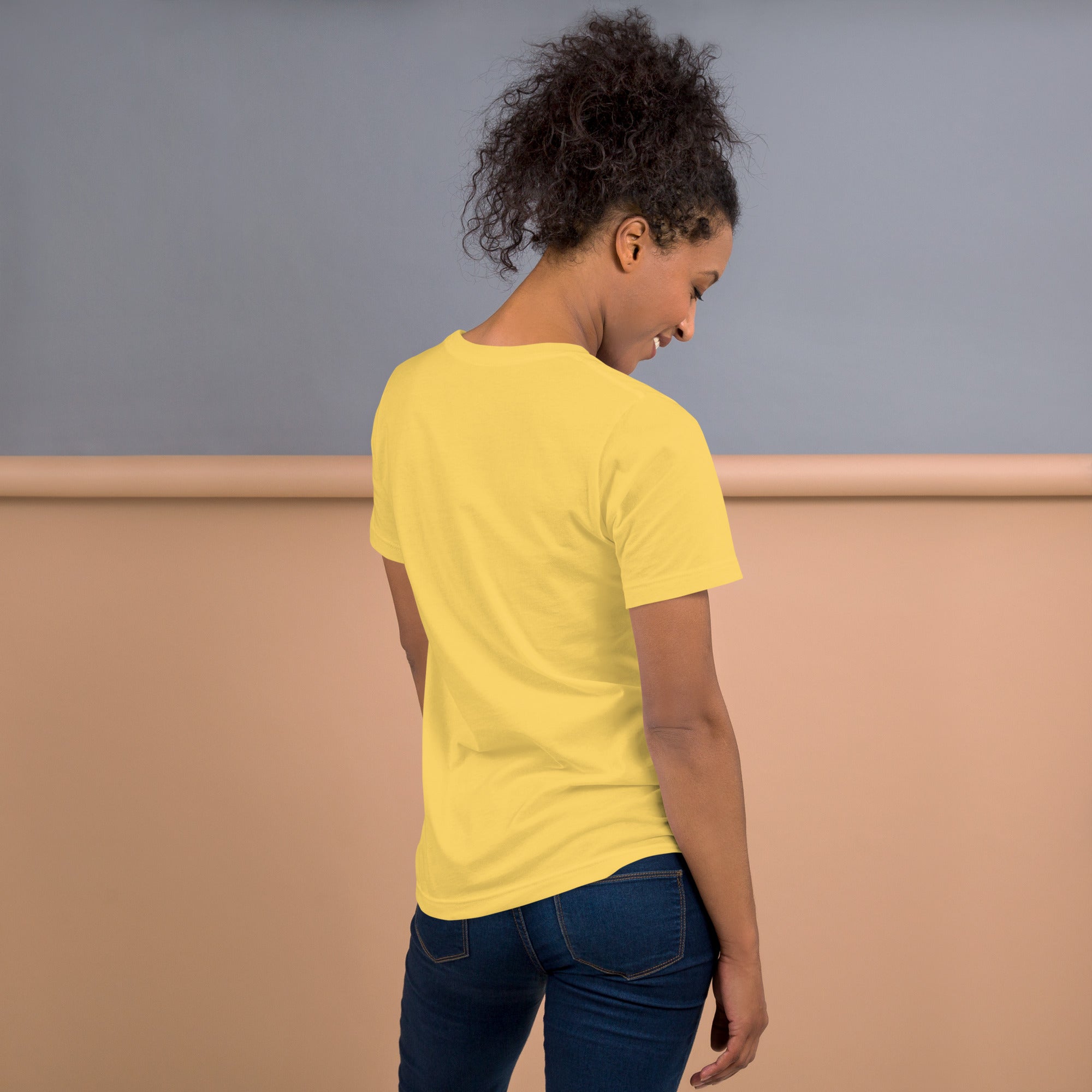 A woman shown from the back wearing a plain yellow short-sleeve crewneck t-shirt and dark jeans, standing in front of a taupe and gray color-blocked studio wall.