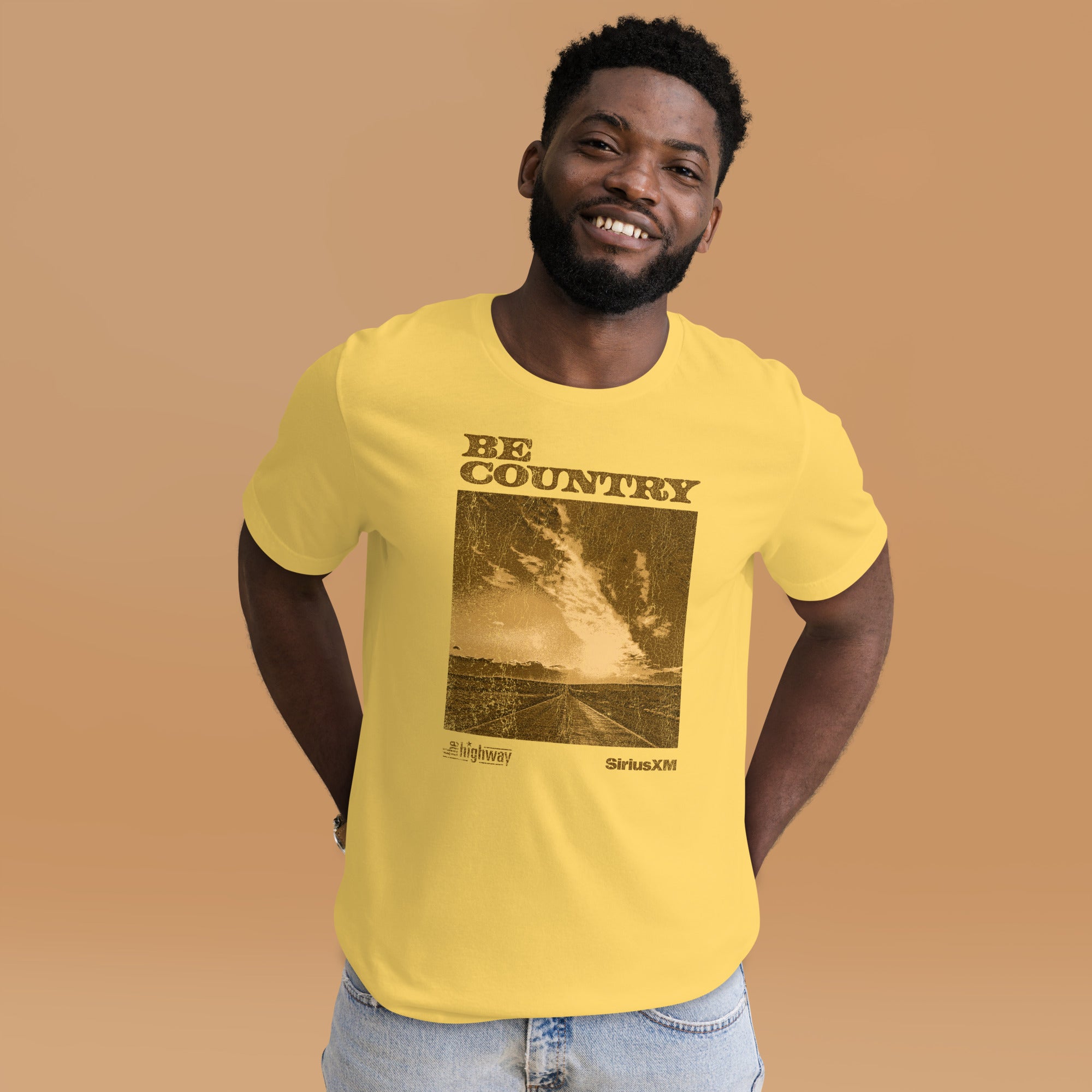 A man wearing a yellow t-shirt with a sepia-toned print on the front; the design features a dramatic rural road and sky photo with distressed texture, topped with the words 'BE COUNTRY' in bold letters, and logos for 'The Highway' and 'SiriusXM' underneath.