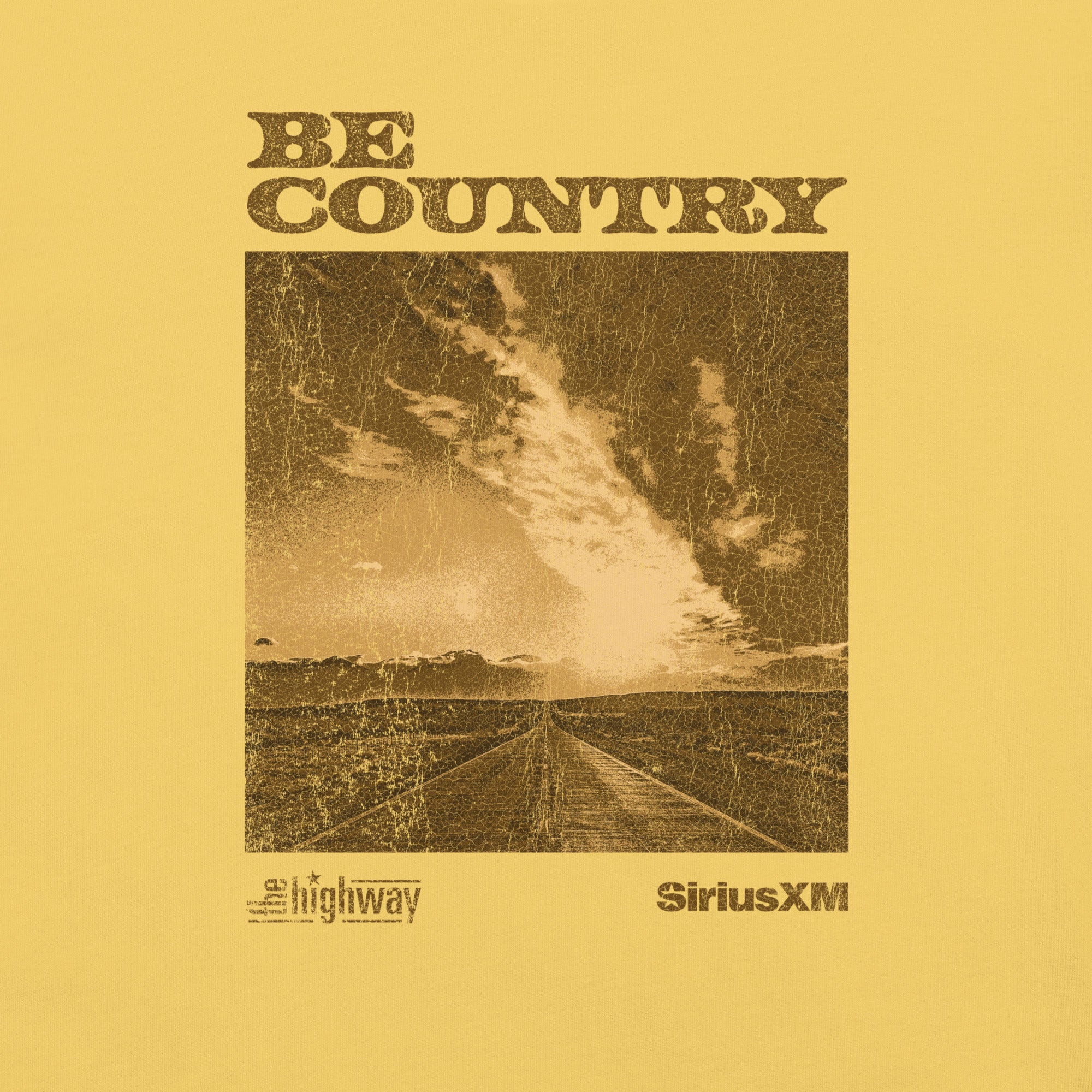 Zoomed-in view of the t-shirt’s graphic design showing a sepia-tinted image of a country road and cloudscape with the words 'BE COUNTRY' in distressed lettering above, and 'The Highway' and 'SiriusXM' logos printed beneath.