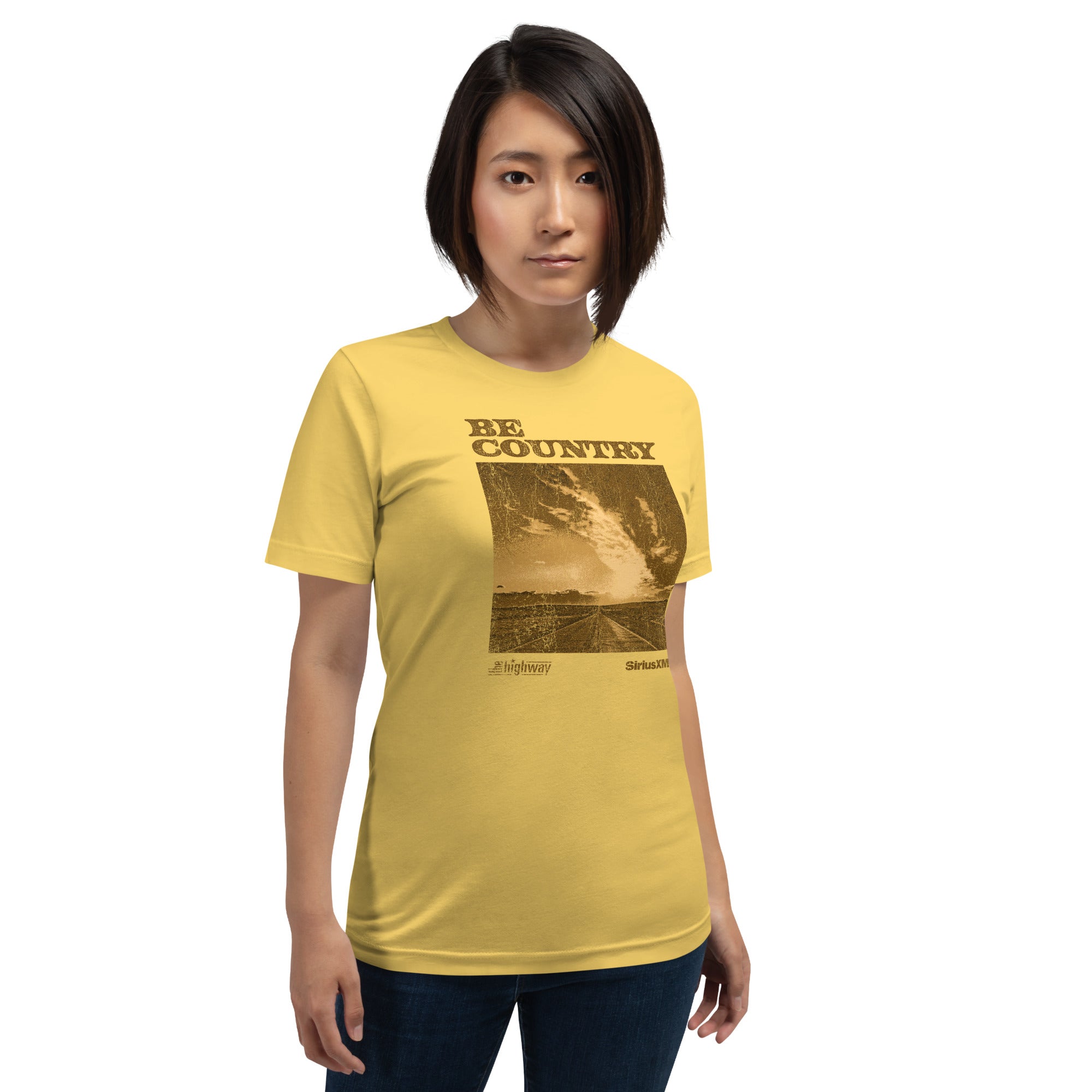 A person wearing a yellow unisex T-shirt with a brown-toned graphic on the front. The graphic shows a rural road leading toward a wide sky filled with streaked clouds. Above the image, the text reads 'BE COUNTRY' in distressed block lettering. Below the image are two logos: 'The Highway' on the left and 'SiriusXM' on the right. The person is standing against a white background and looking forward.