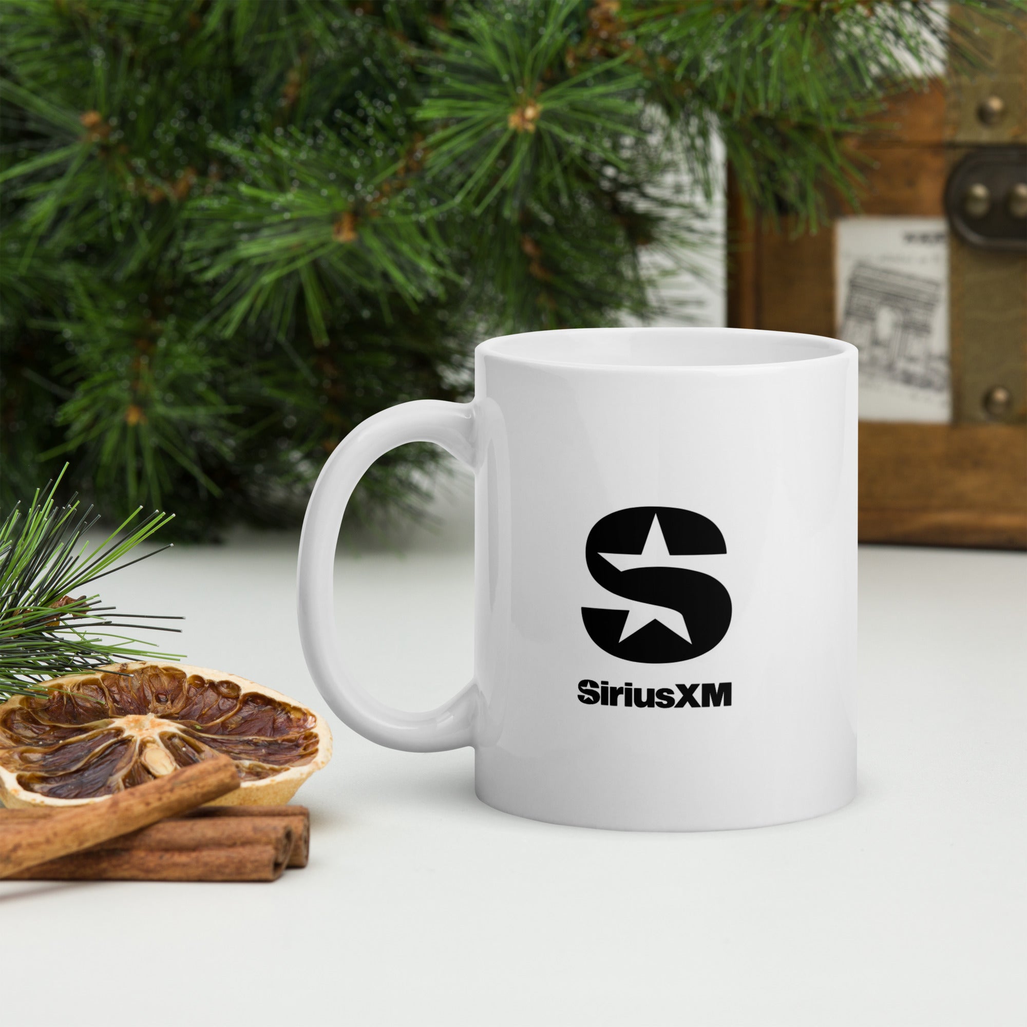 White ceramic mug with the black ‘SiriusXM’ logo featuring a stylized star inside the letter ‘S,’ shown on a holiday-themed background with pine branches, dried citrus slices, and cinnamon sticks.
