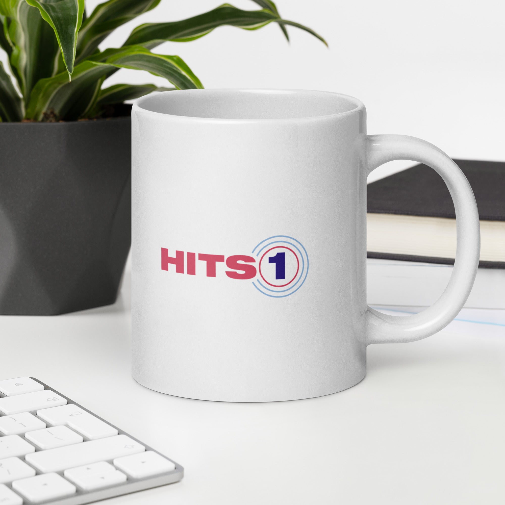 White mug with 'HITS 1' logo on a desk with a plant and books in the background